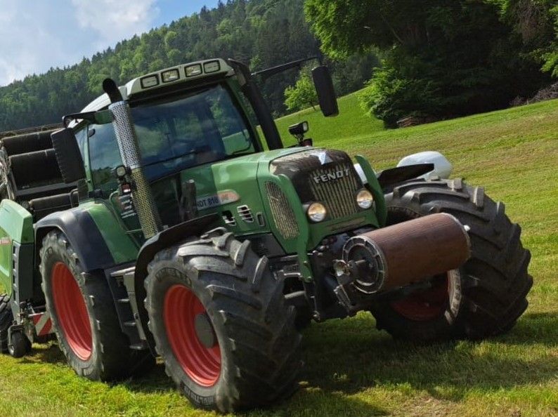 Grüner Traktor auf einem grasbewachsenen Hügel; große Reifen, brauner Zylinder an der Vorderseite, Bäume im Hintergrund.