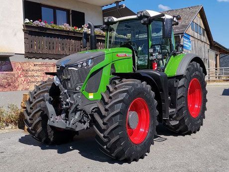 Ein grüner und grauer Traktor mit roten Rädern parkt vor einem Holzgebäude.