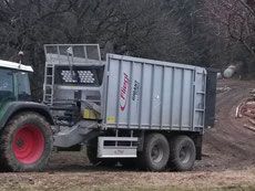 Traktor, der einen silbernen Industrieanhänger auf einer unbefestigten Straße zieht, wahrscheinlich für die landwirtschaftliche Nutzung.