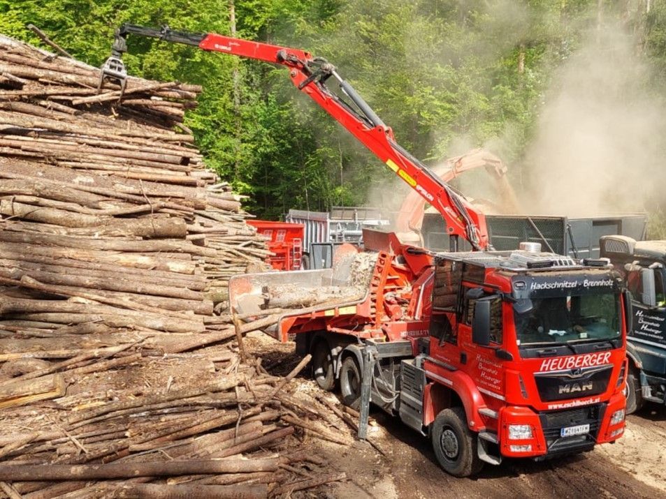 Roter LKW mit Kran lädt Baumstämme auf einem Holzplatz, im Hintergrund Bäume und Holzspäne.