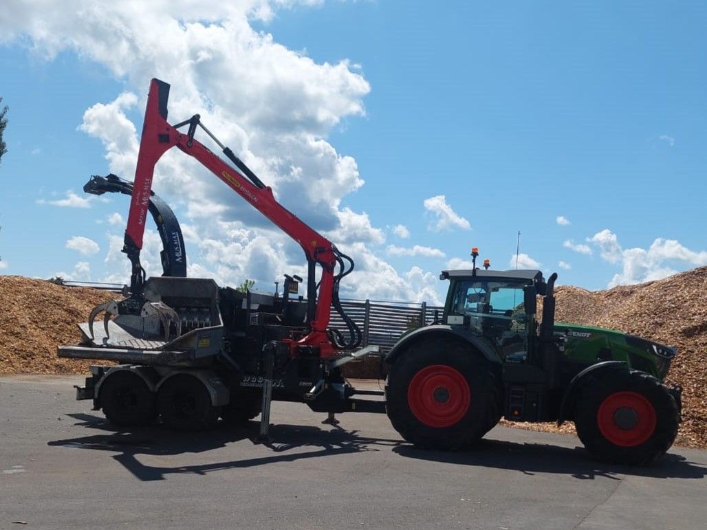 Traktor mit rotem Kran lädt an einem sonnigen Tag mit blauem Himmel und Wolken Baumstämme auf einem Holzplatz.