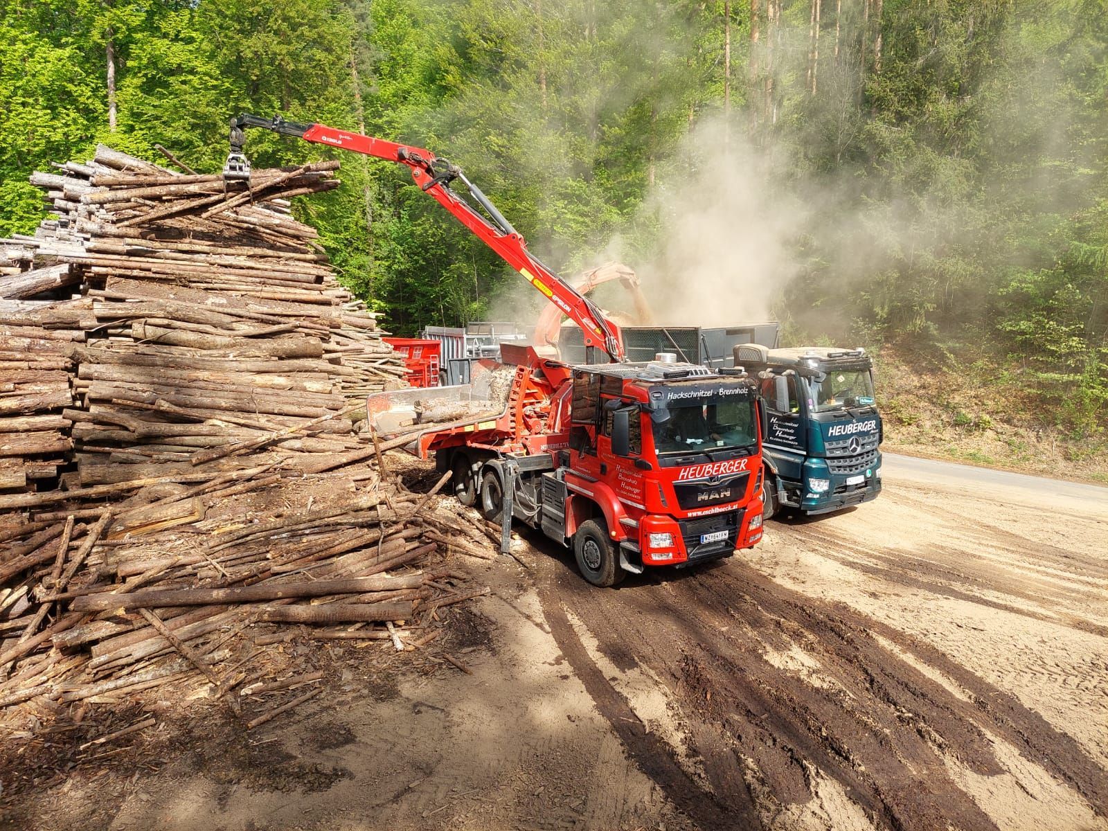 Ein Holztransporter mit rotem Kran belädt einen dunklen LKW mit Holzspänen; im Hintergrund ein staubiger Wald.
