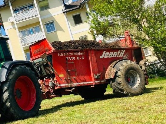 Roter Miststreuer, der an einem Traktor auf einer Wiese befestigt ist; im Hintergrund ein Gebäude.