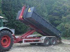 Traktor, der einen angehobenen, blauen Kippanhänger zieht; im Freien, auf einer Schotterfläche.