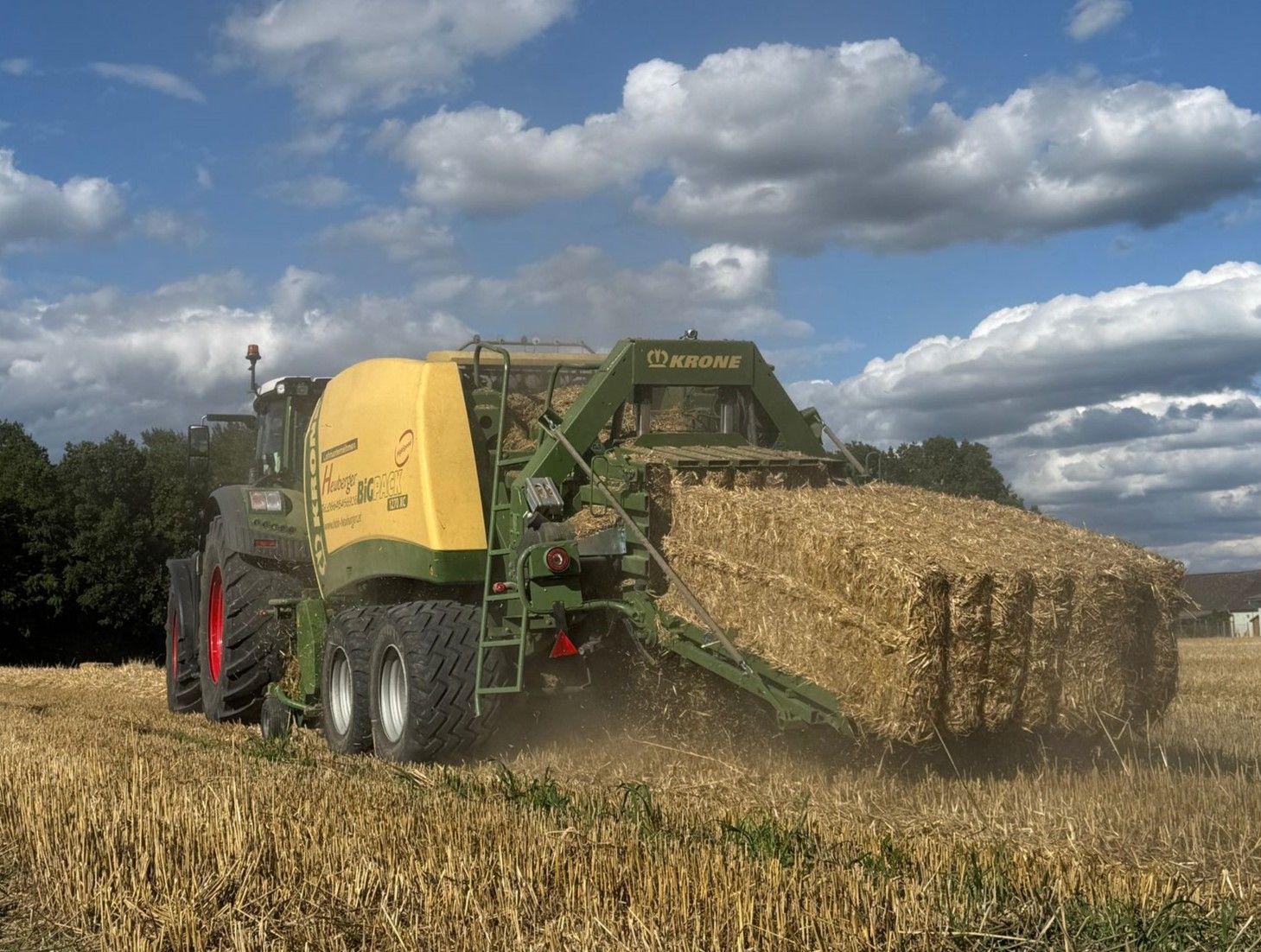Eine von einem Traktor gezogene Ballenpresse formt unter bewölktem Himmel auf einem Stoppelfeld einen großen, rechteckigen Ballen.