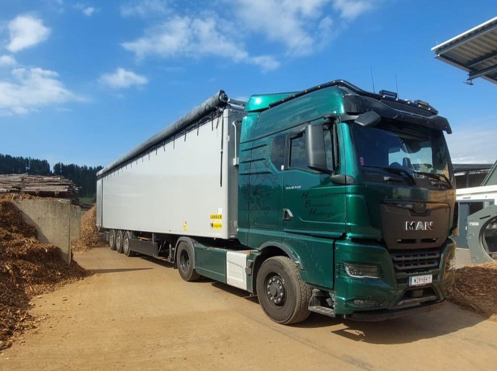 Grüner Sattelschlepper mit weißem Anhänger auf einem Feldweg neben Holzspänen, unter blauem Himmel.