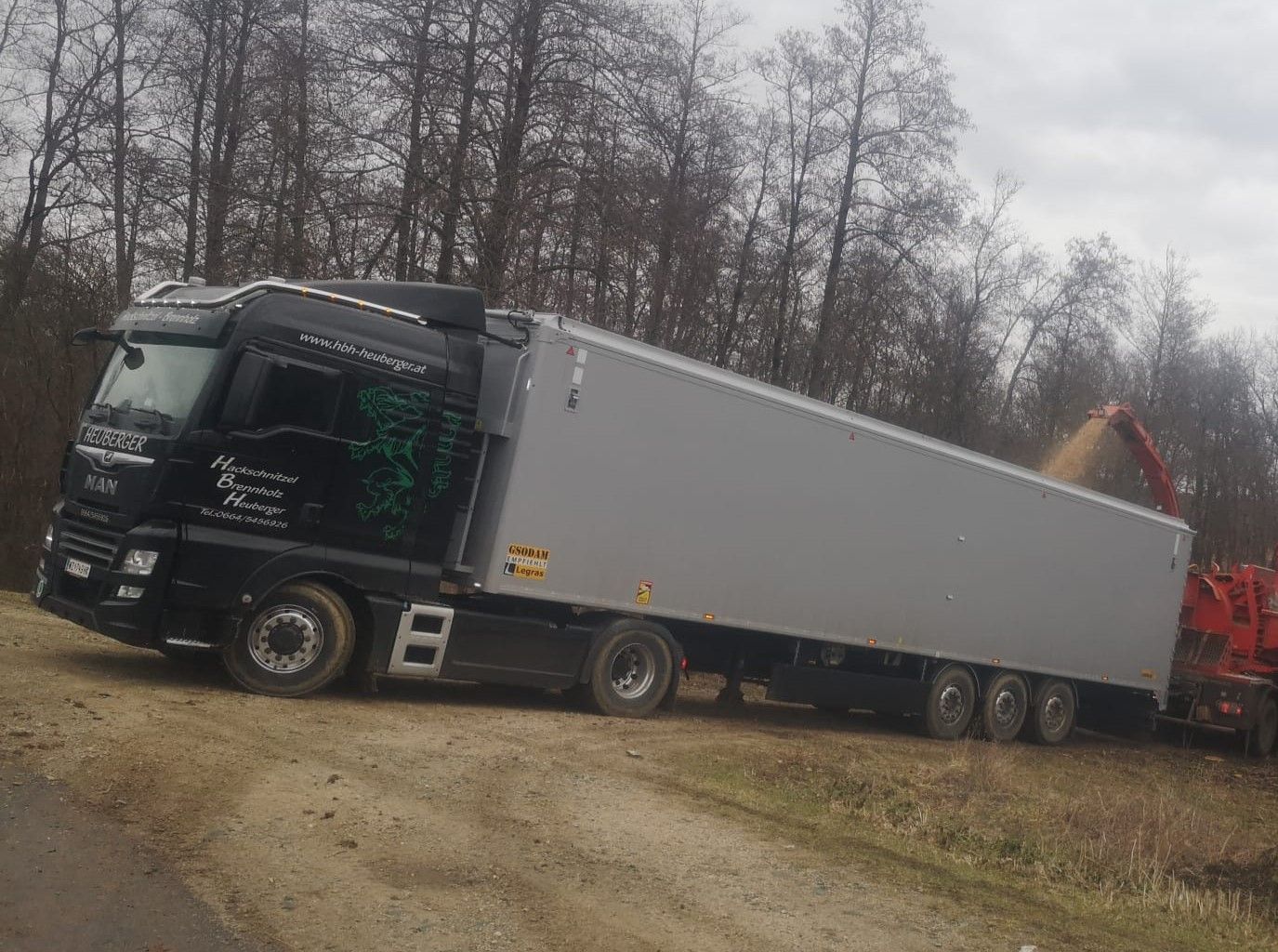Schwarzer Sattelschlepper mit grauem Anhänger, der auf einer unbefestigten Straße in der Nähe eines Waldes geparkt ist und mit Holzspänen beladen wird.
