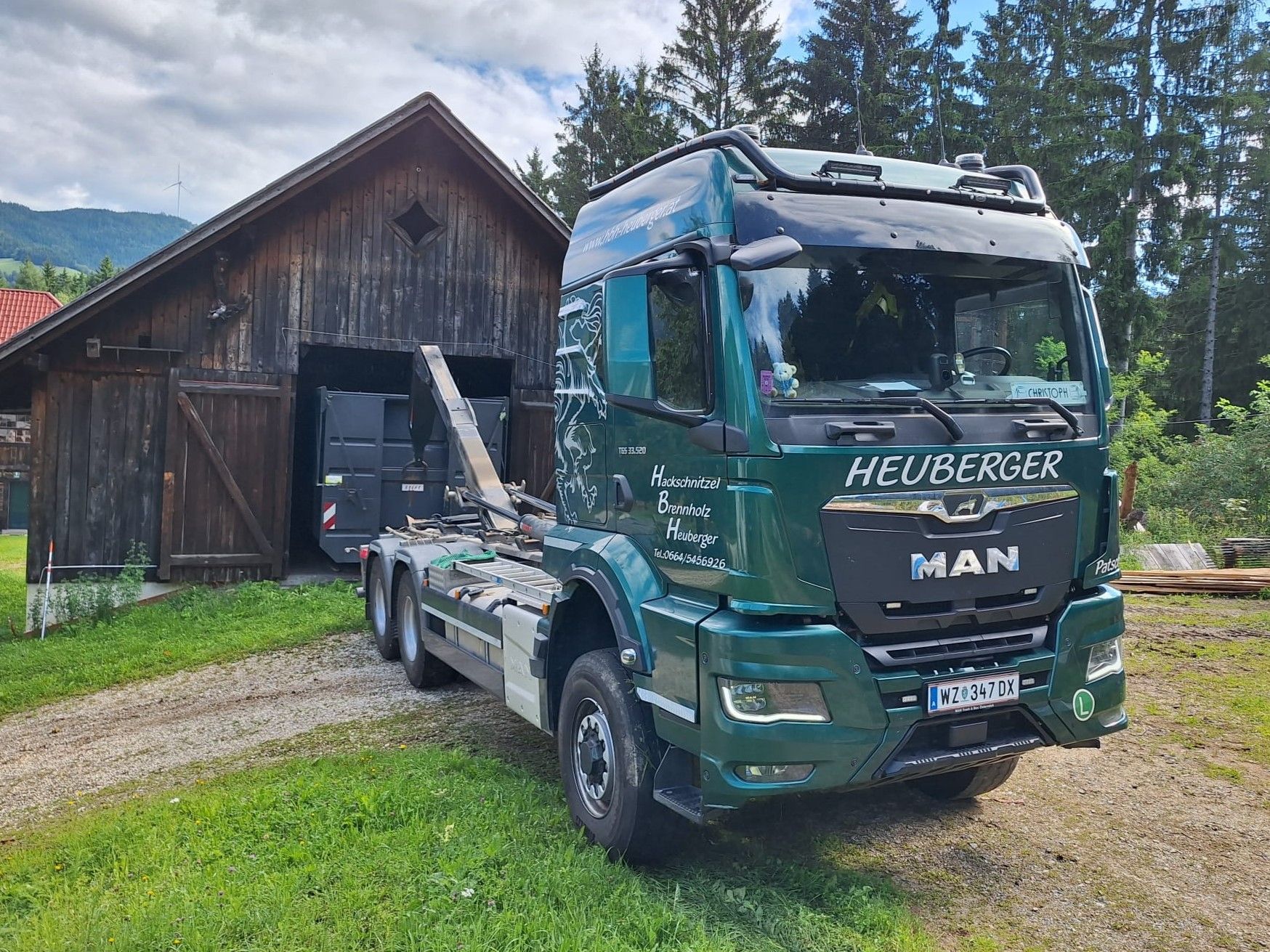 Grüner MAN-LKW mit Container, geparkt vor einer Holzscheune.