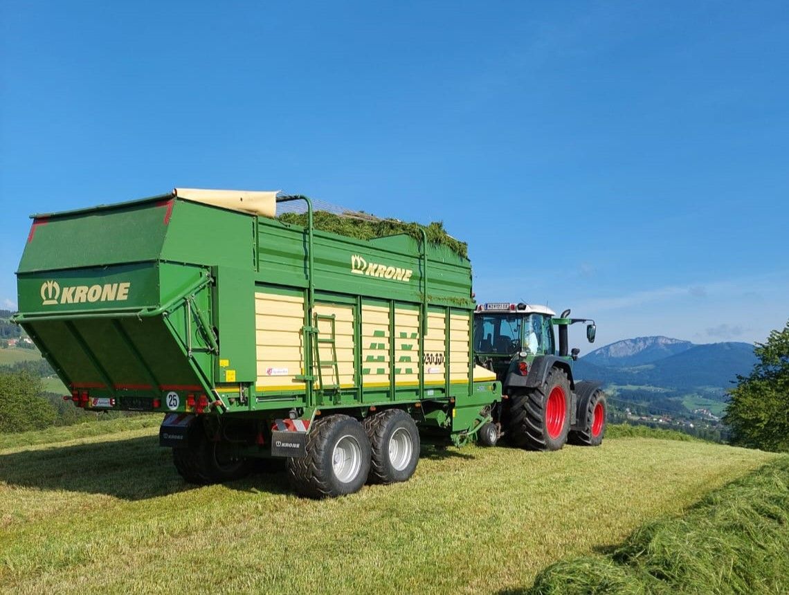 Grüner Traktor zieht einen Krone-Ladewagen auf einem Feld unter blauem Himmel.