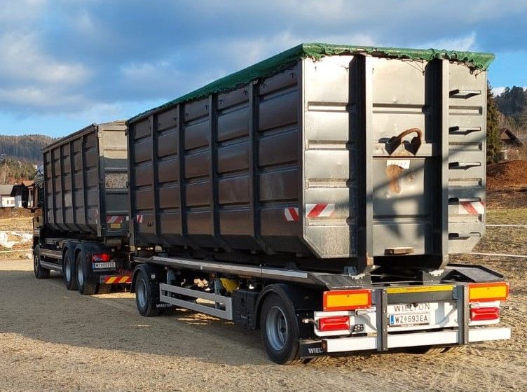 Ein dunkelgrauer Sattelzug mit zwei großen, abgedeckten Containern parkt an einem sonnigen Tag im Freien.