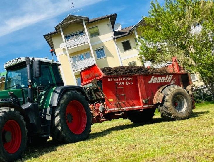 Grüner Traktor zieht einen roten Miststreuer auf einem Grashang in der Nähe eines beigen Gebäudes unter blauem Himmel.