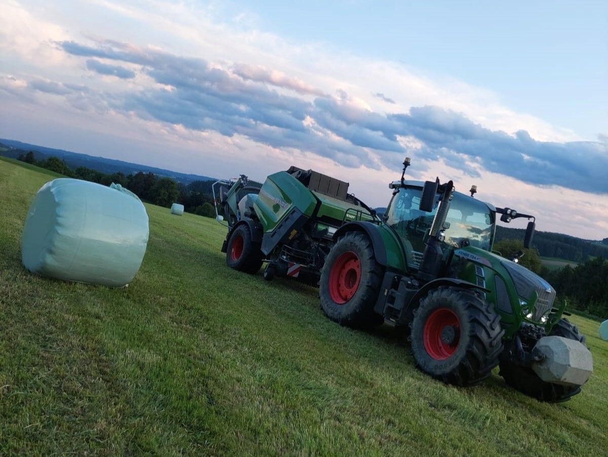 Grüner Traktor presst Heu auf einem grasbewachsenen Hügel; im Vordergrund eingewickelter Heuballen.