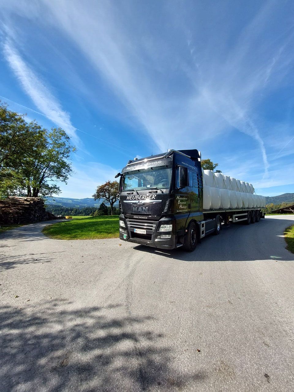 Schwarzer Sattelschlepper auf einer asphaltierten Straße, transportiert weiße Heu- oder Siloballen, blauer Himmel, Bäume und eine bergige Landschaft.