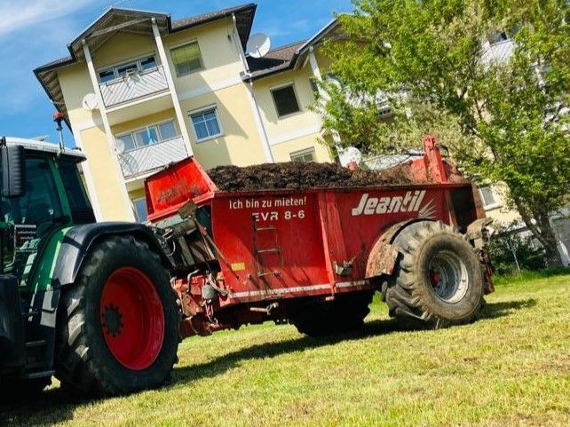 Ein roter Traktor mit einem Anhänger voller Mist auf einer Rasenfläche in der Nähe eines gelben Gebäudes.