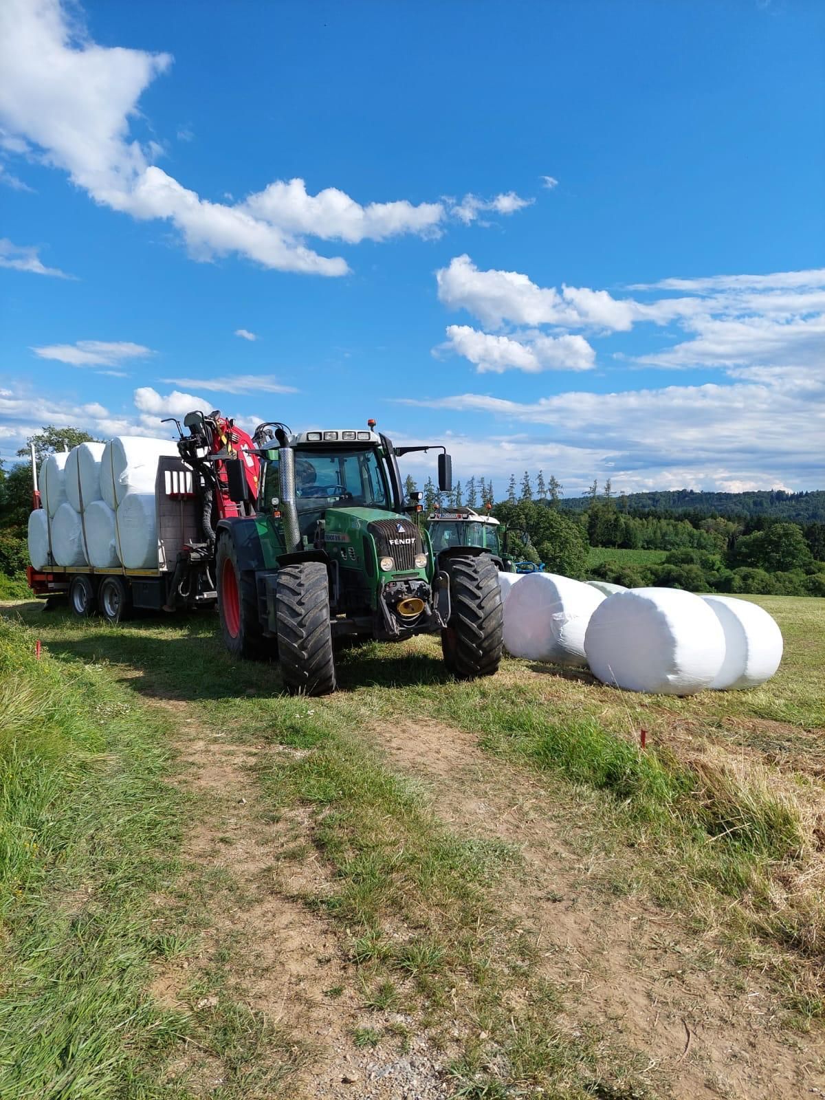 Traktor transportiert Heuballen auf einem Anhänger auf einem Feld unter blauem Himmel mit Wolken.