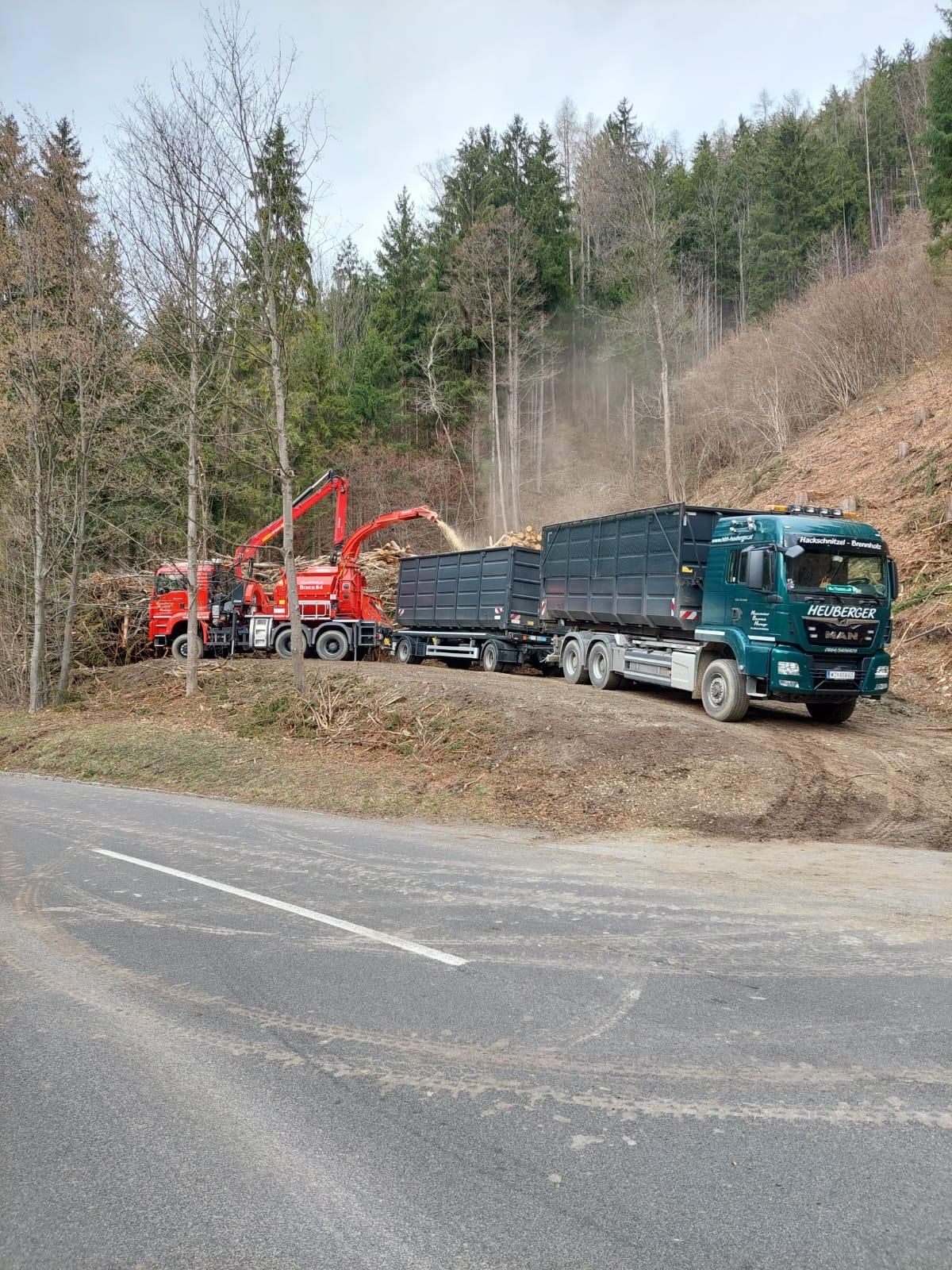 Ein roter Holzhäcksler und ein grüner LKW-Anhänger parken neben einer Straße in einem Wald.