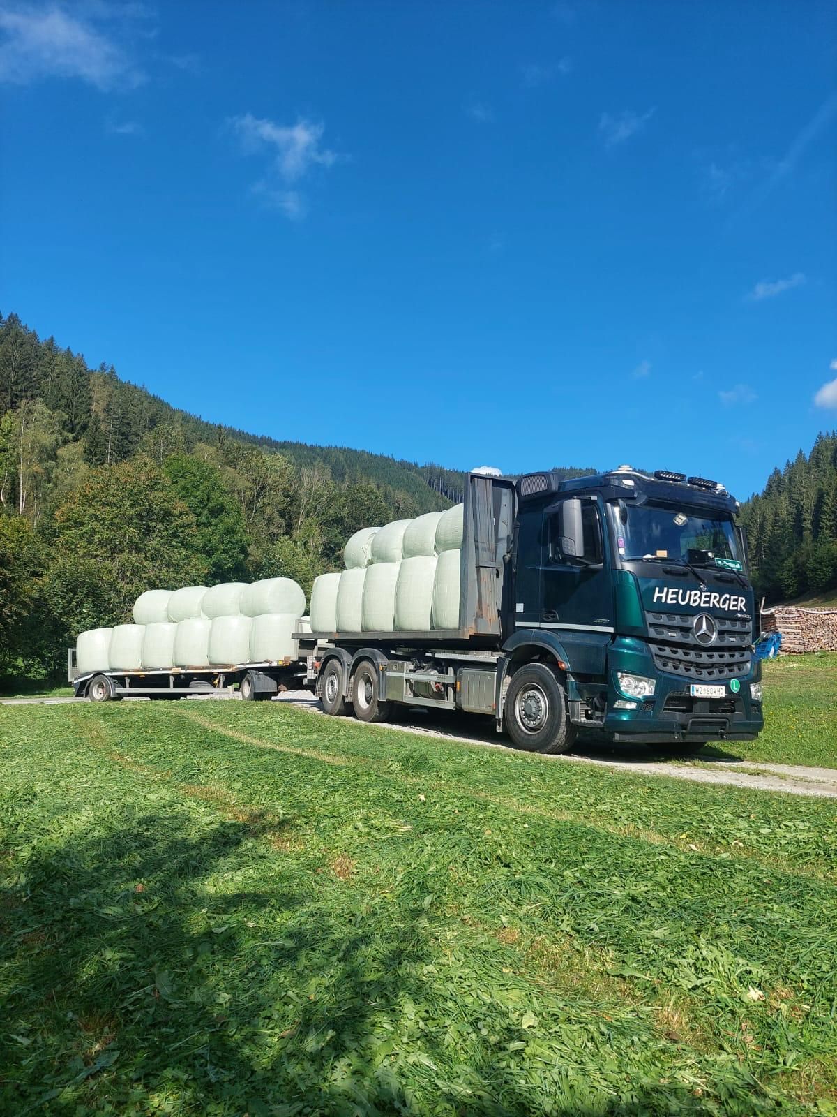 Ein LKW mit Anhänger, beladen mit Heuballen, auf einer Wiese, im Hintergrund Bäume und blauer Himmel.