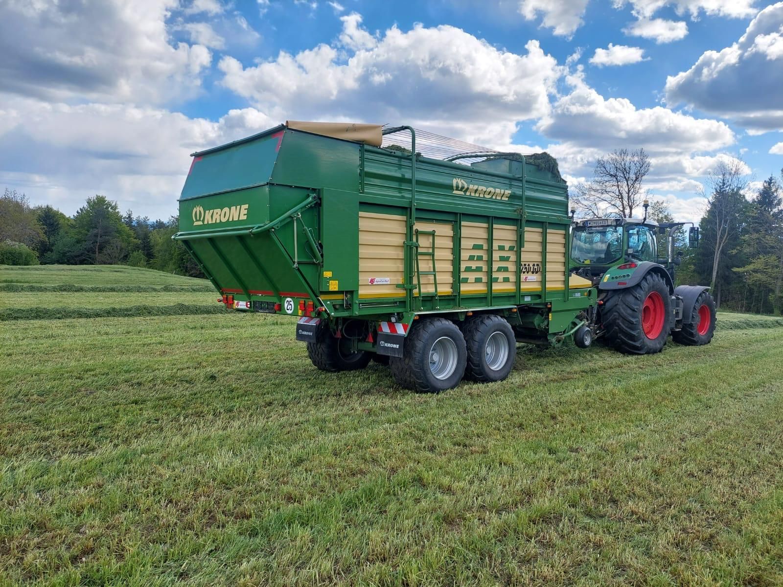 Ein grüner Krone-Futterwagen, gezogen von einem blauen Traktor, auf einem Feld unter bewölktem Himmel.