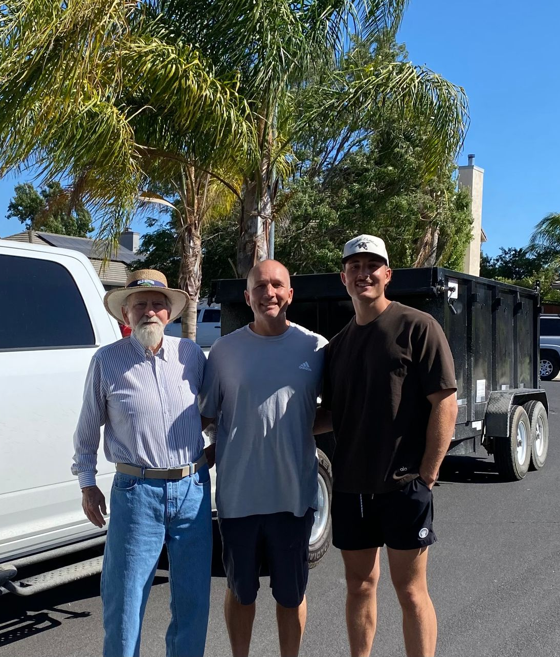 Three men standing next to a truck and a trailer. One man is wearing a cowboy hat, the others have on caps. Sunny outdoor setting.