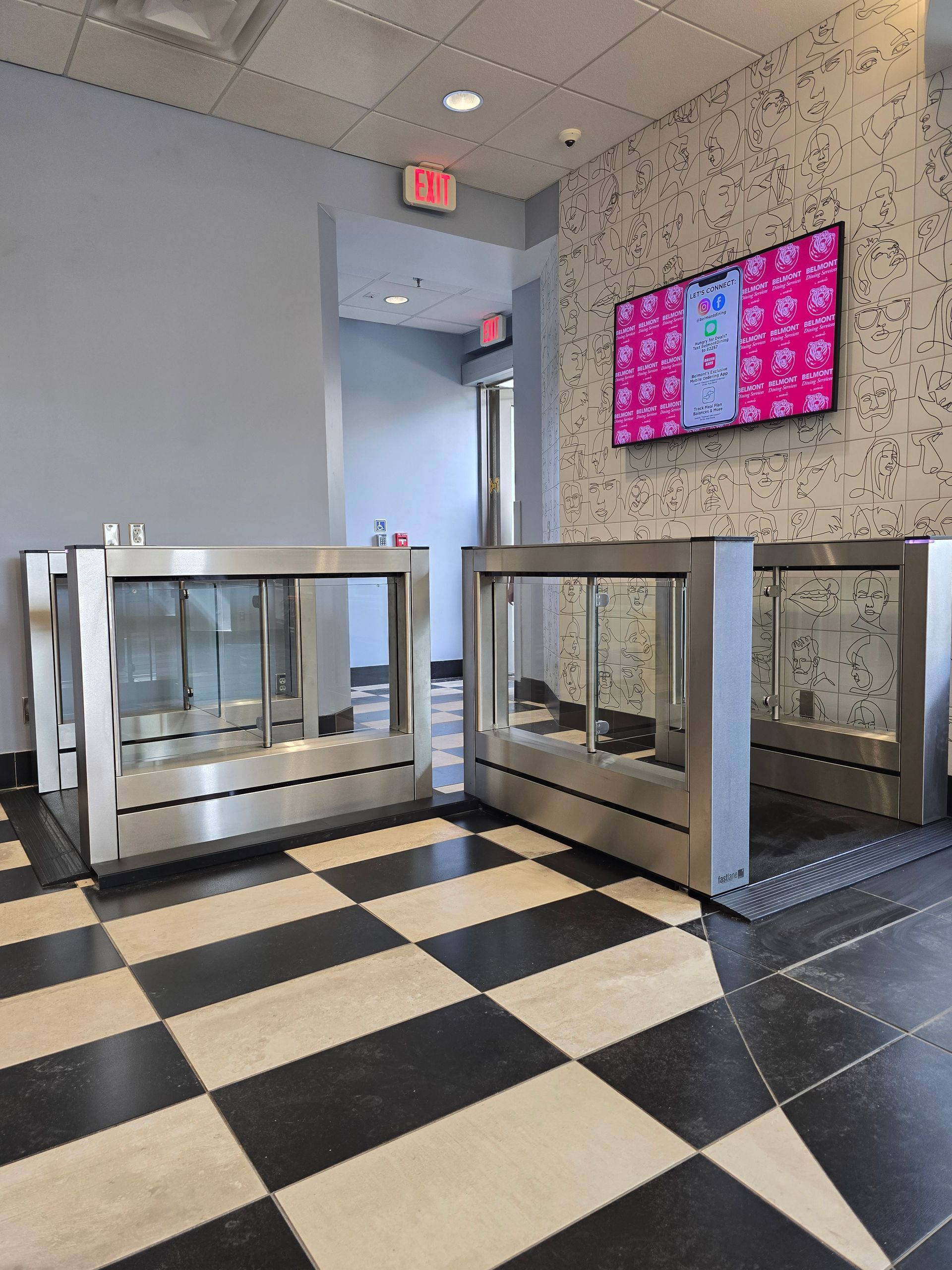 Stainless steel turnstiles in a lobby with a checkered floor and a television on the wall.