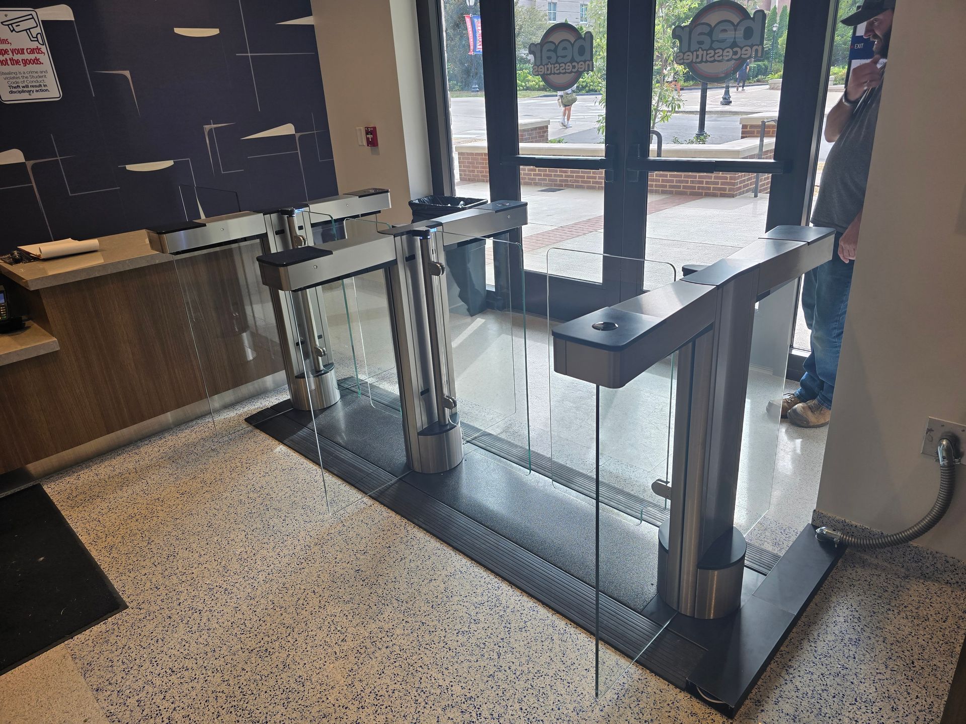 Entrance with glass security turnstiles and a person in a mask visible.