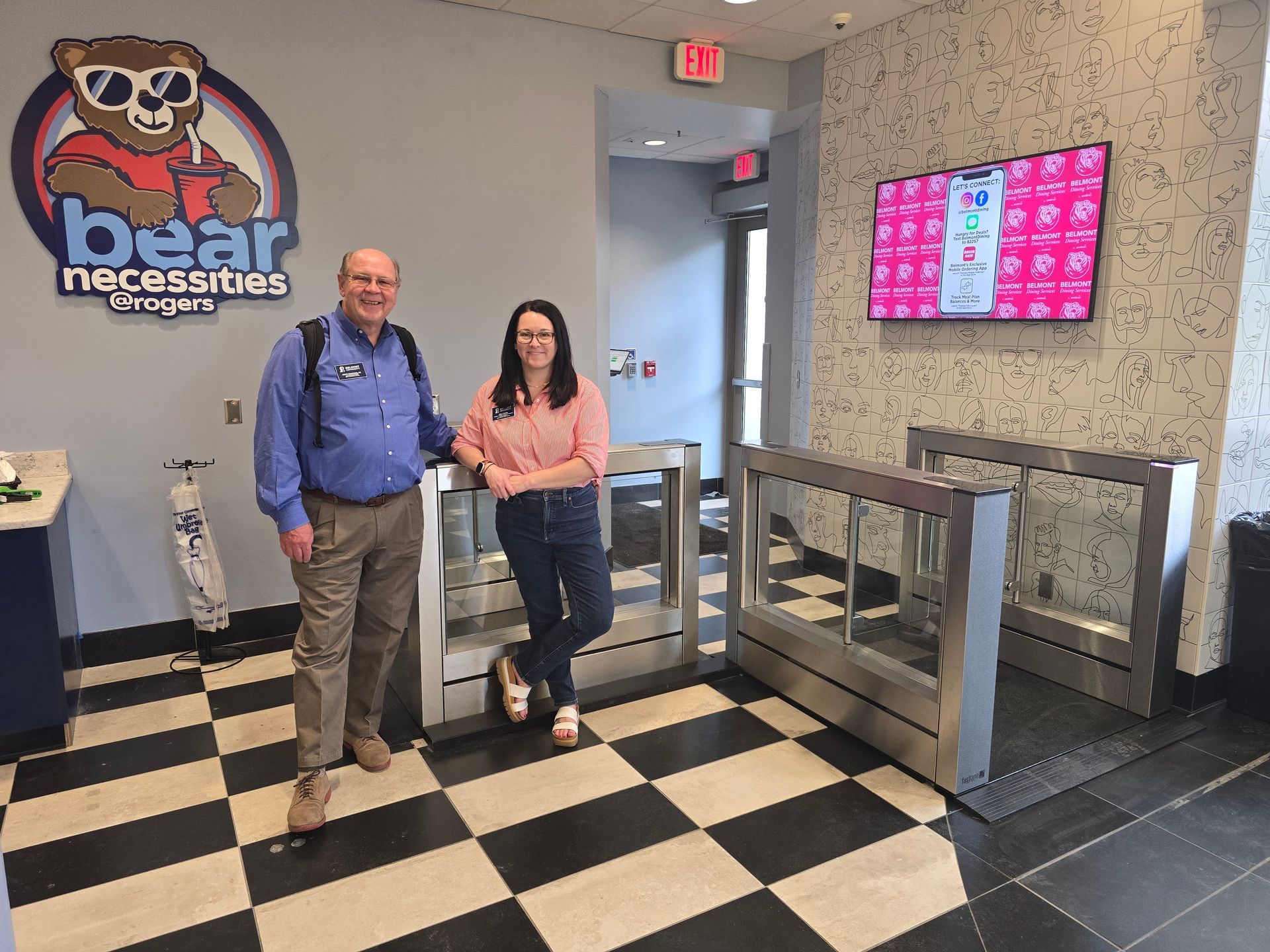 Two people stand near a security turnstile inside a store with a black and white tiled floor.