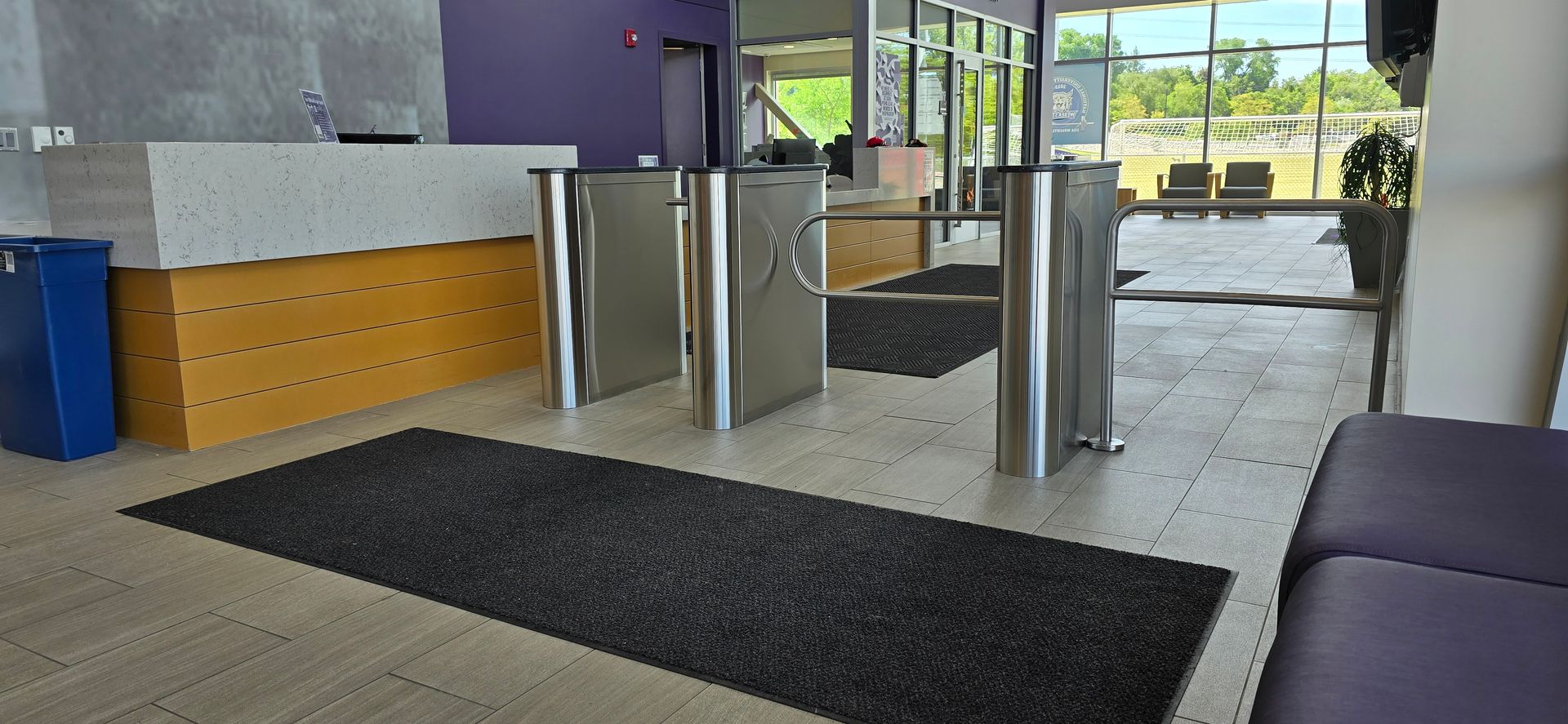 Turnstiles in an entrance lobby with a purple accent wall and windows overlooking an outdoor area.