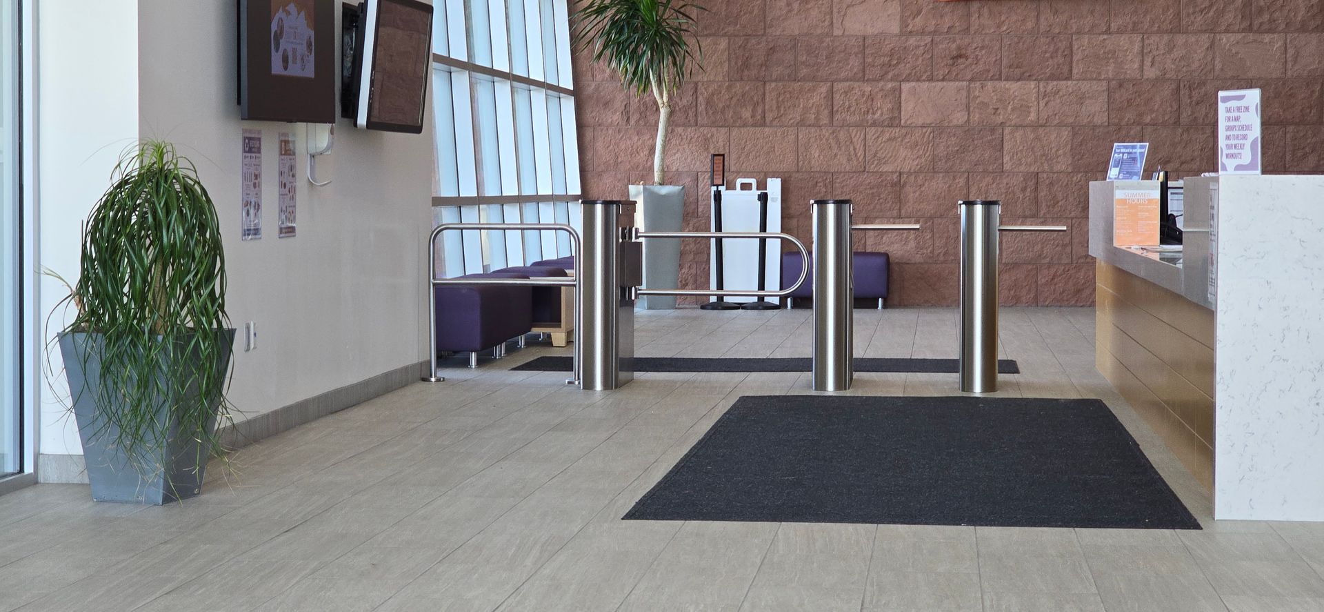 Lobby with turnstiles, reception desk, potted plant. Brick wall background, dark doormat.