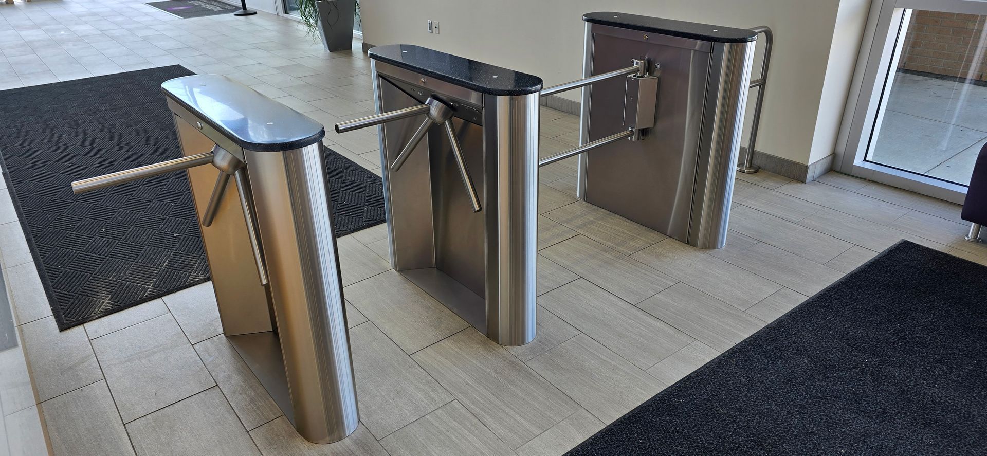 Turnstiles in a building entrance, stainless steel finish, with black mats in front.