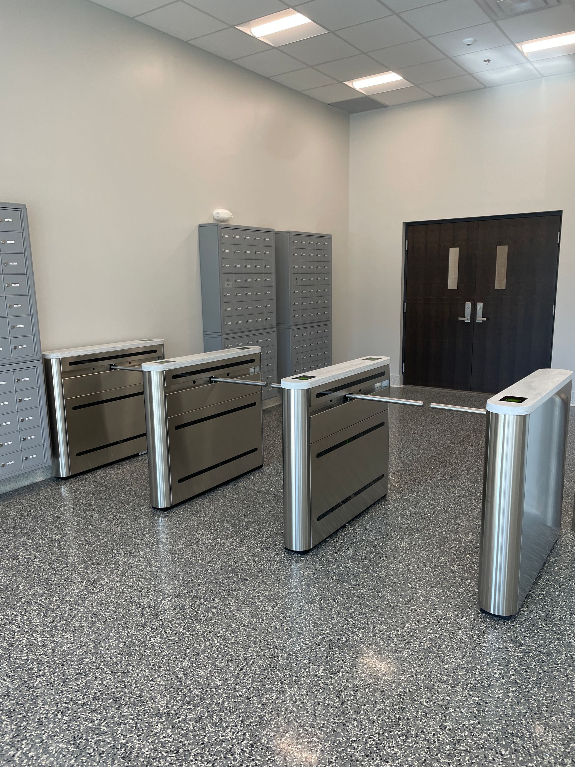 Turnstiles and lockers in a modern entryway with a dark double door at the back.