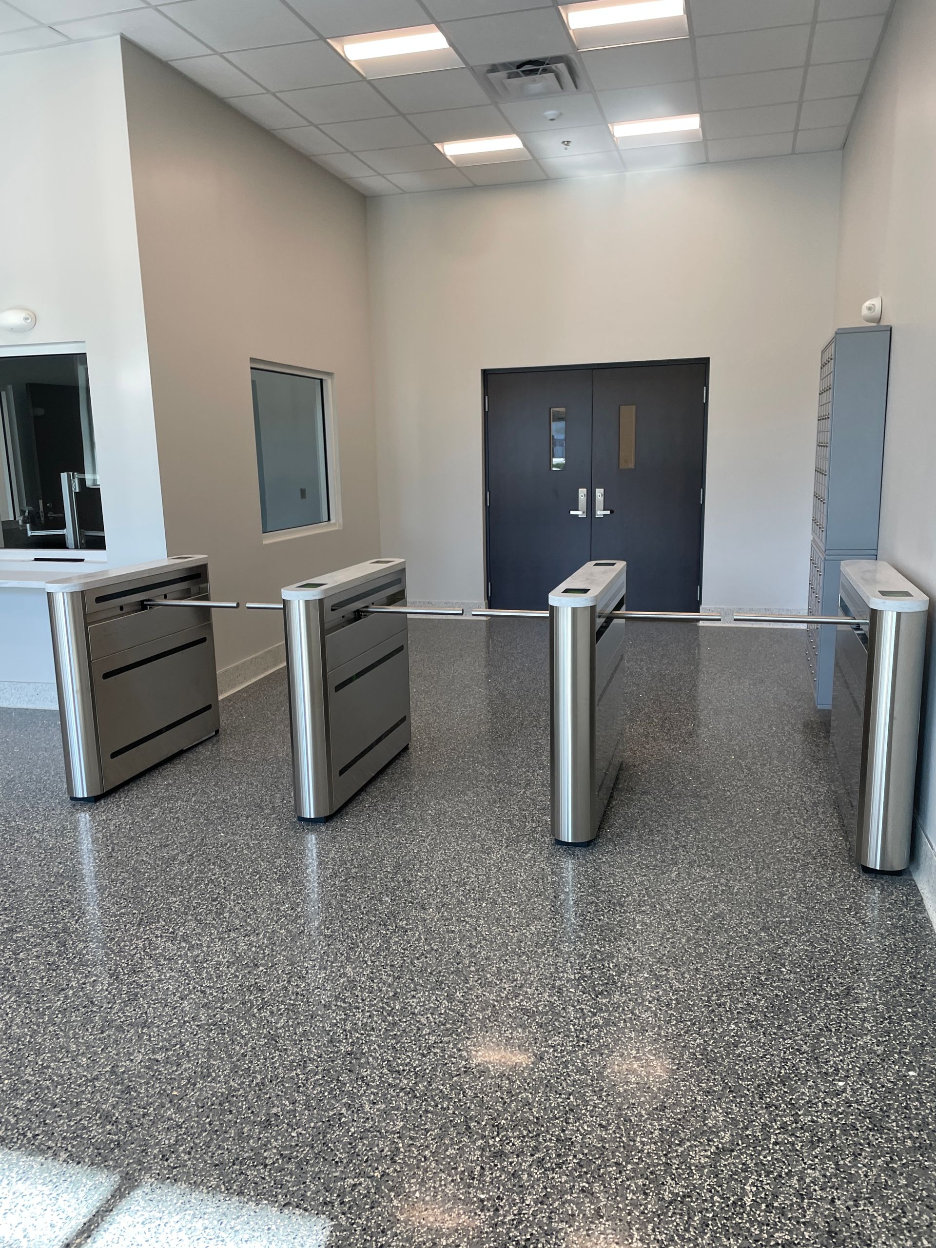 Turnstiles in an entranceway with a dark gray door and speckled gray flooring.