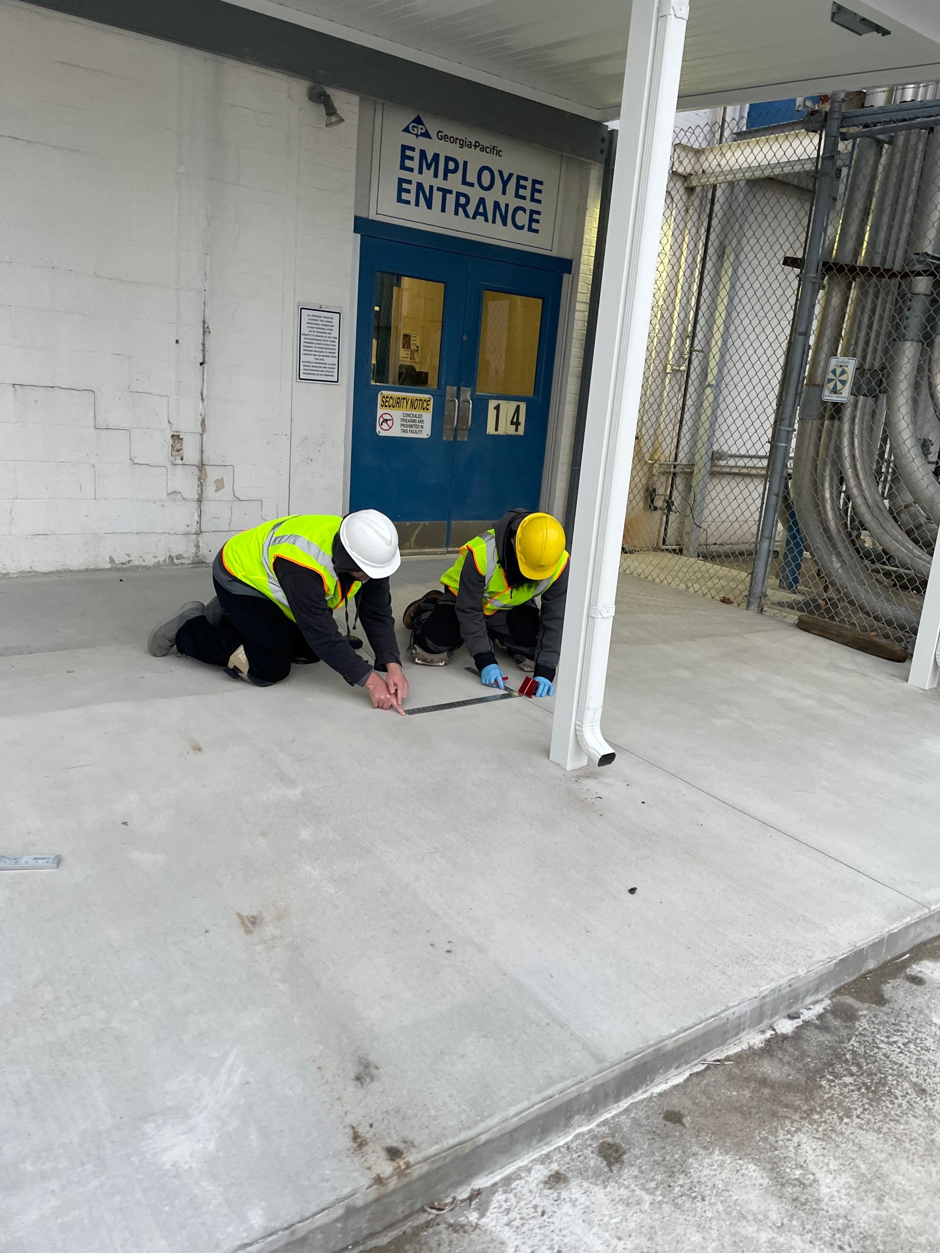 Two workers in safety vests kneeling, working on concrete outside an employee entrance.