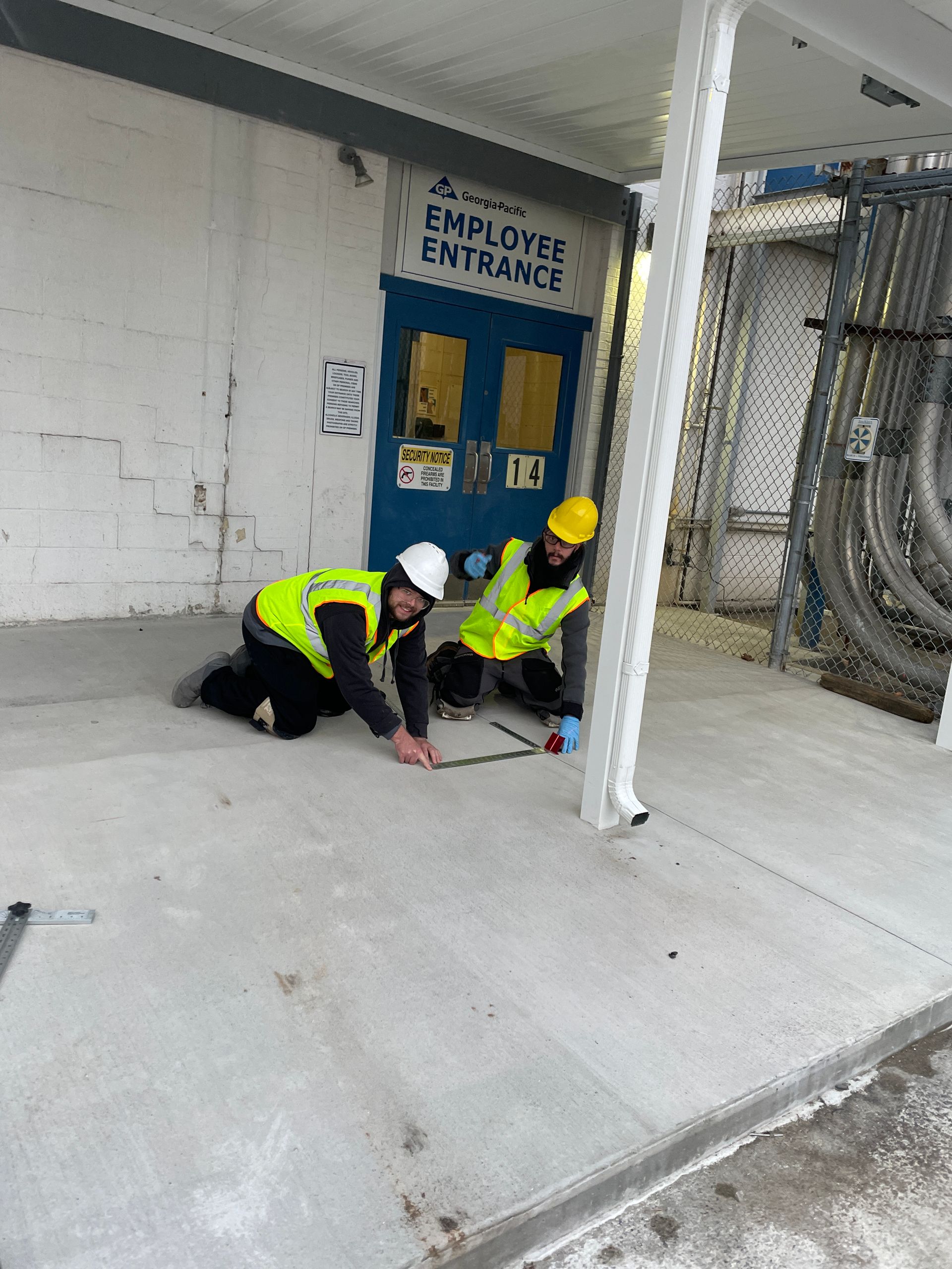 Two workers, wearing hard hats and safety vests, kneeling on concrete by a building entrance.