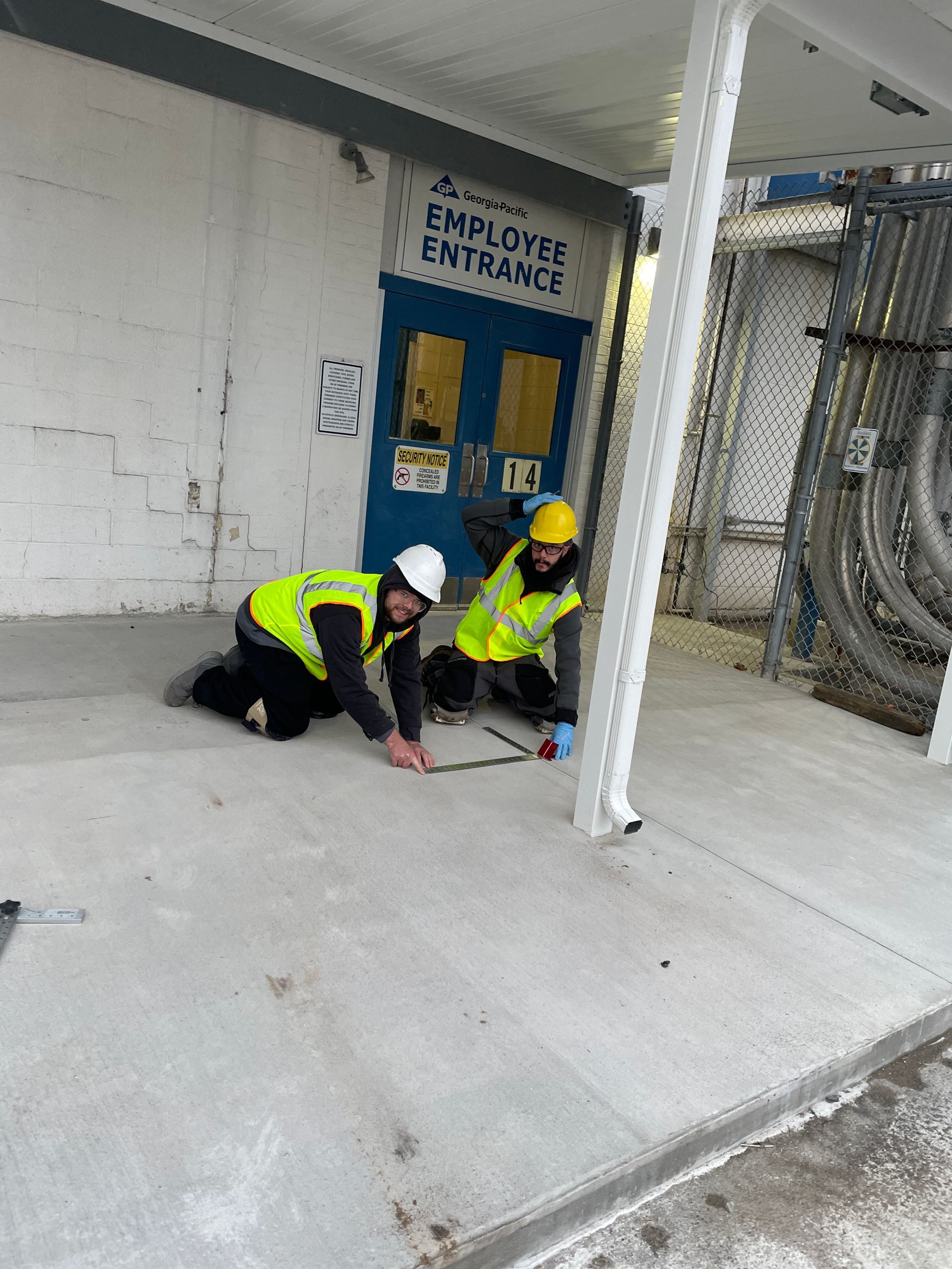 Two workers in safety vests kneeling, examining a concrete surface near an employee entrance.