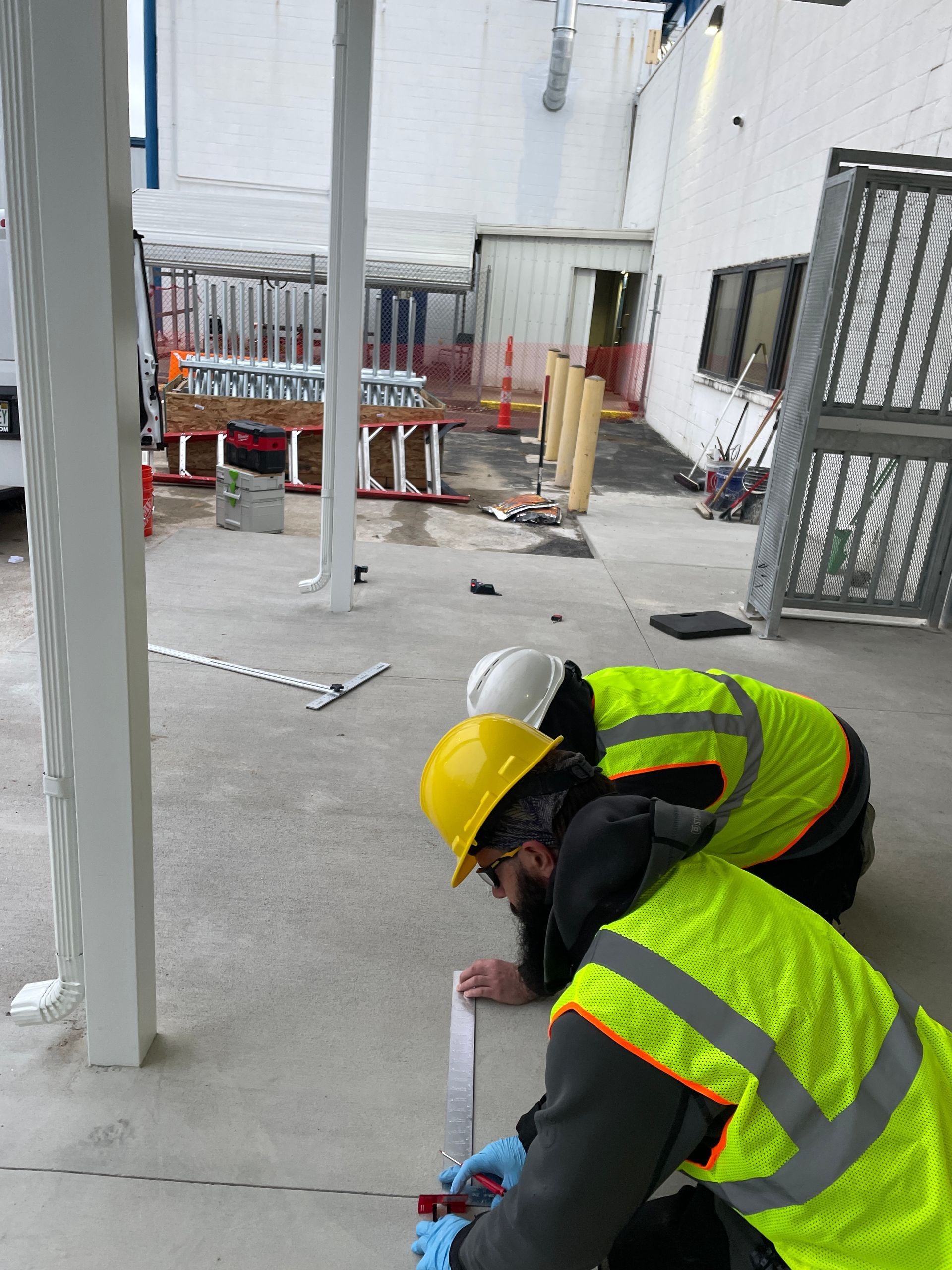 Two construction workers in yellow vests and hard hats measure on a concrete surface.