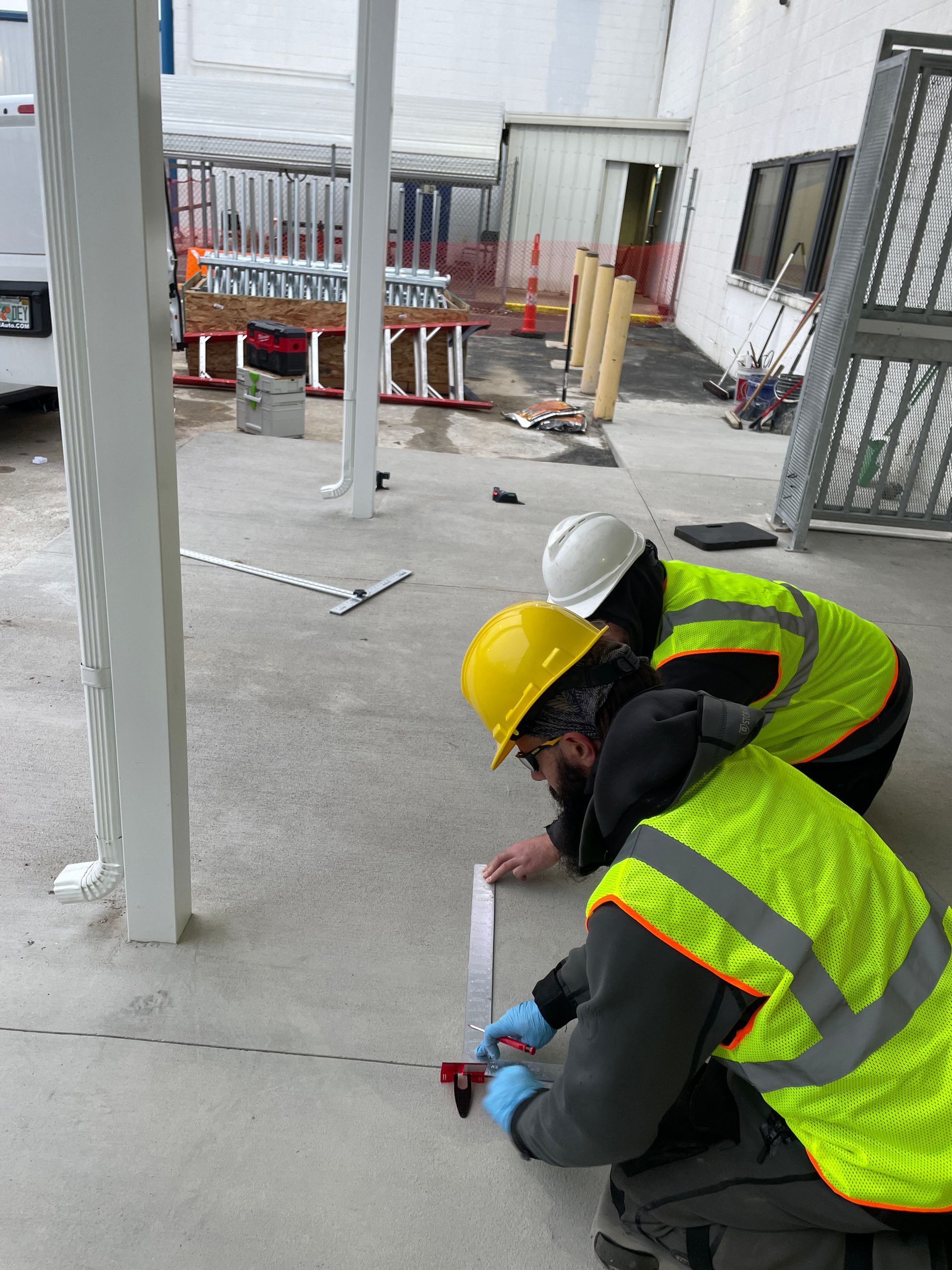 Two workers in safety vests measure concrete; one kneels with a measuring tape and the other observes near a building.