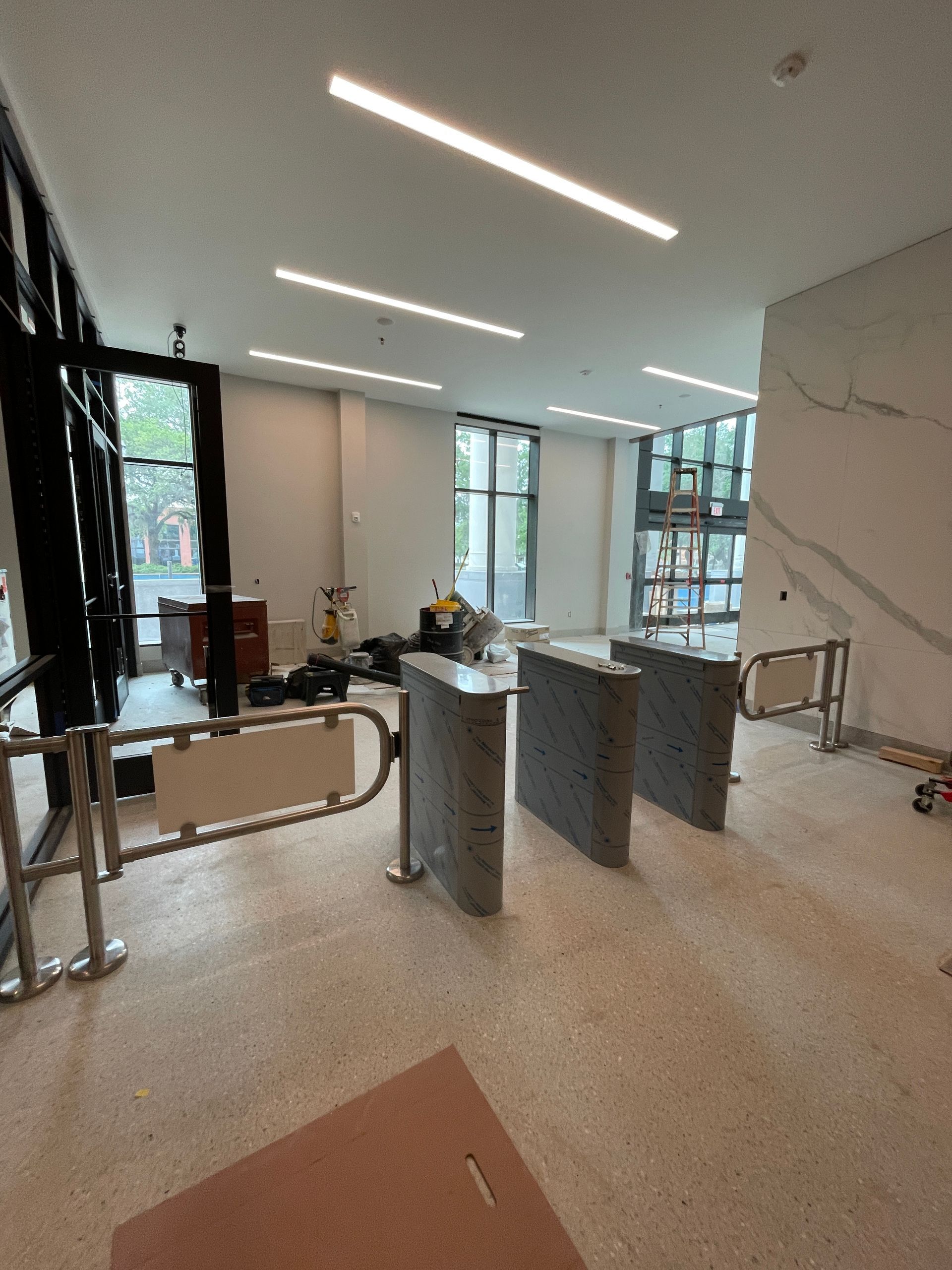 Interior with security turnstiles, large windows, and overhead lights; unfinished construction.