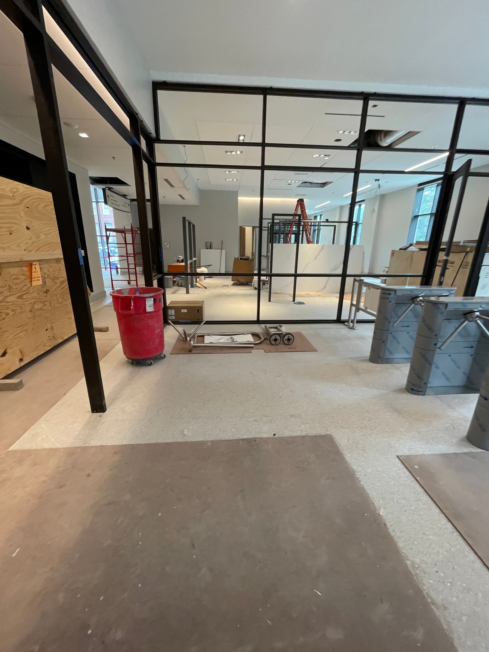 Interior of a construction site with glass walls, concrete floors, red trash bin, and tools.