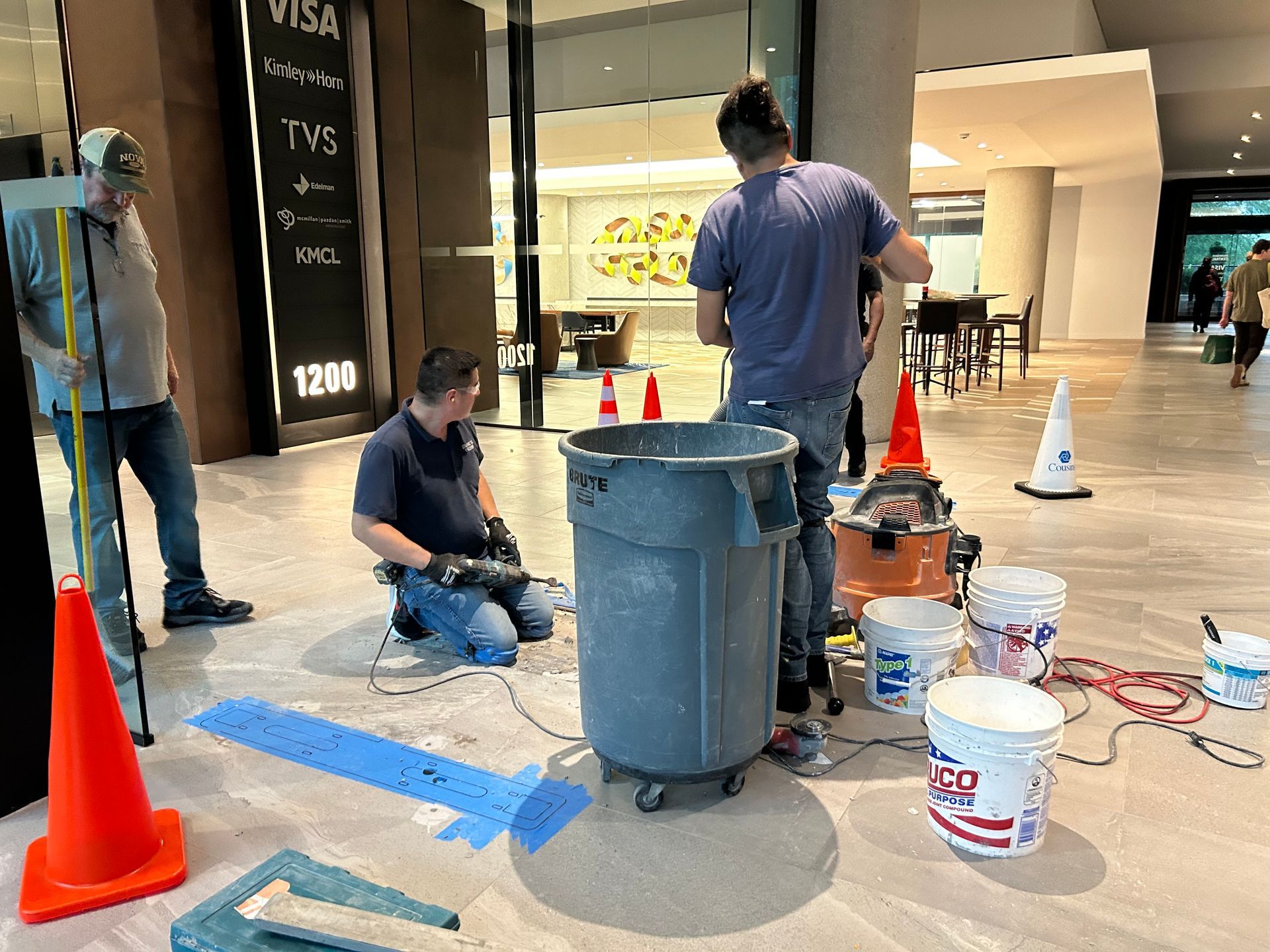 Workers cleaning or repairing flooring in a brightly lit indoor space, using tools, buckets, and a trash can.