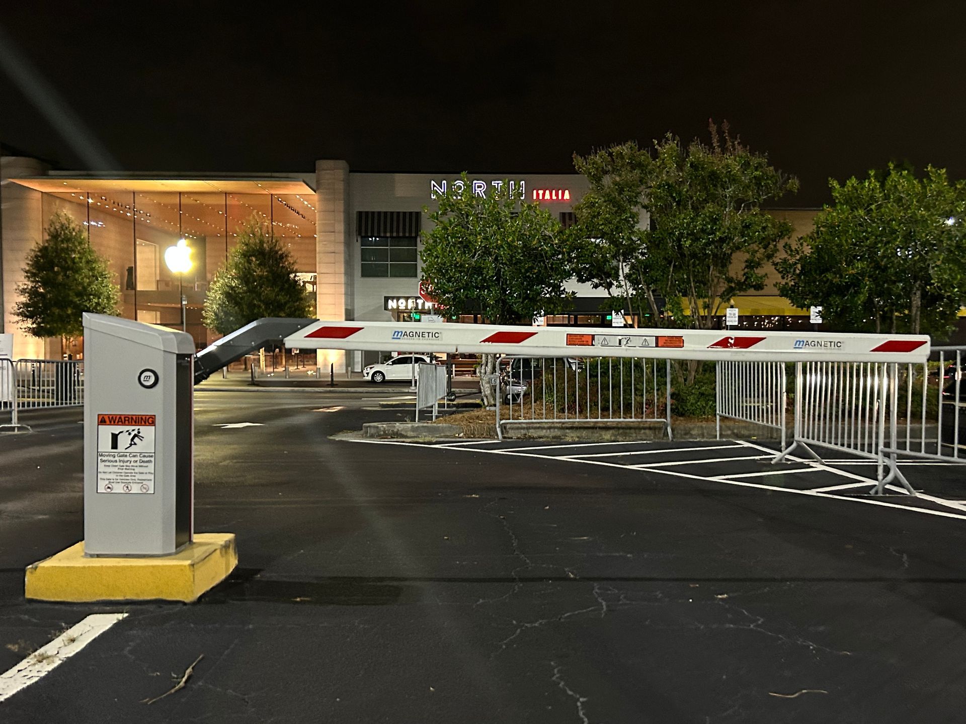 Parking lot entrance with an automatic barrier gate in front of a shopping center at night.