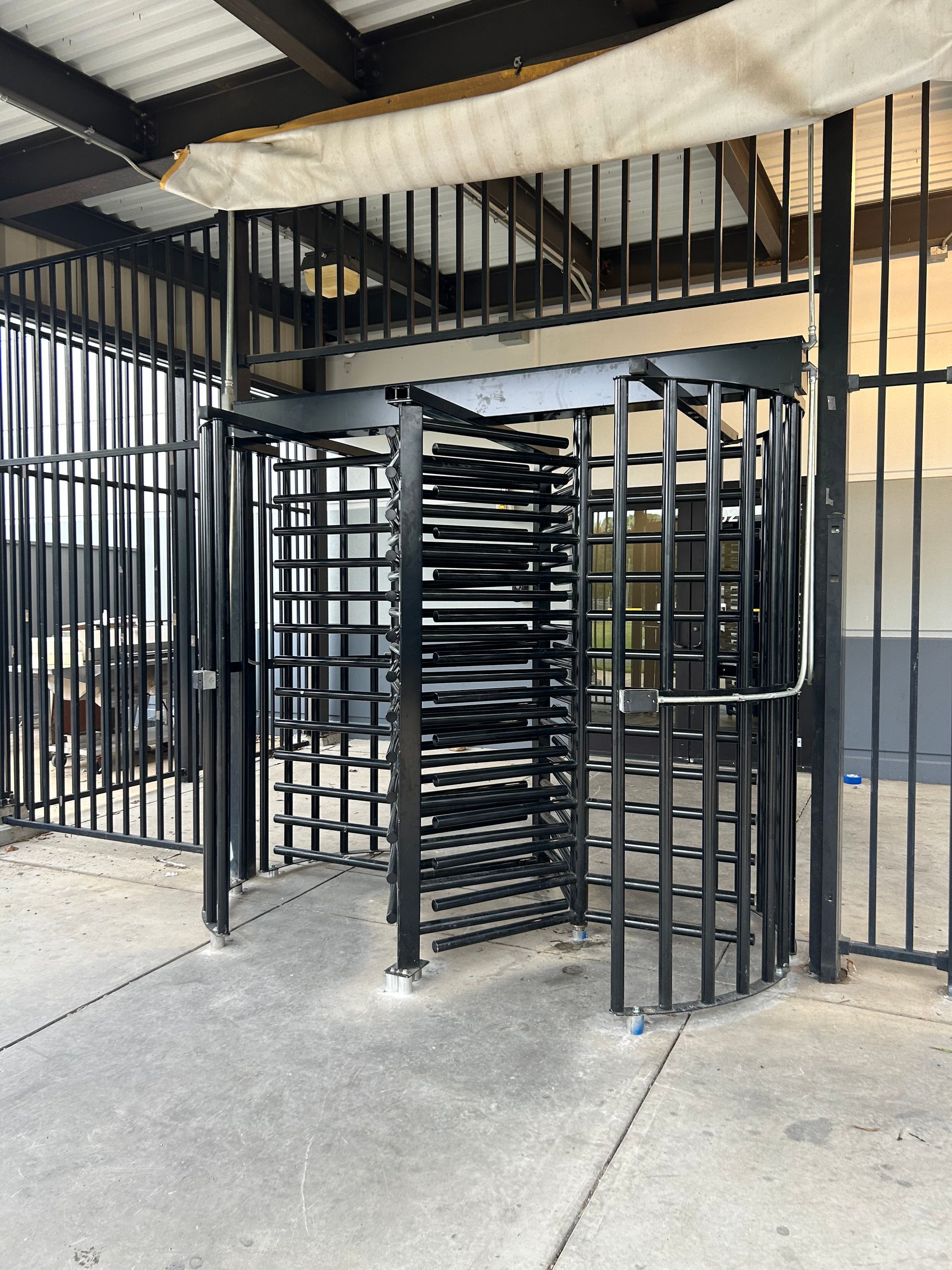 Black metal turnstile in an outdoor area, surrounded by similar metal fencing under a canopy.