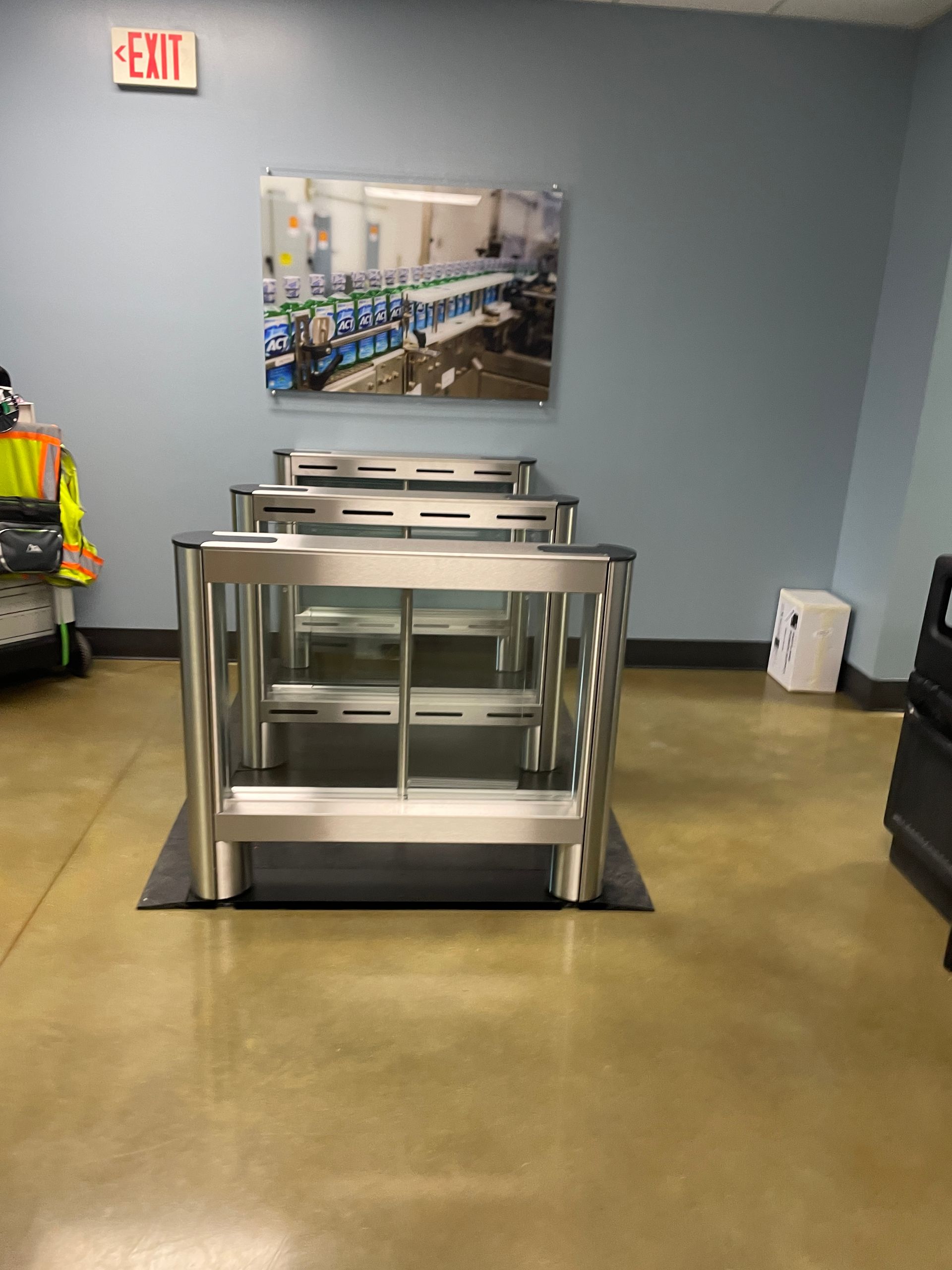 Turnstiles in front of a blue wall with a photo, an exit sign, and a concrete floor.