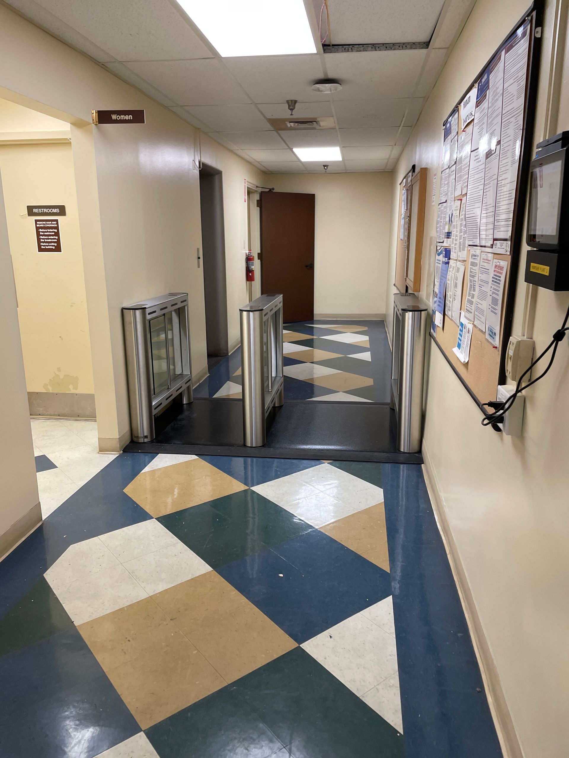 Hallway with security turnstiles, bulletin board, patterned floor, and a closed door.