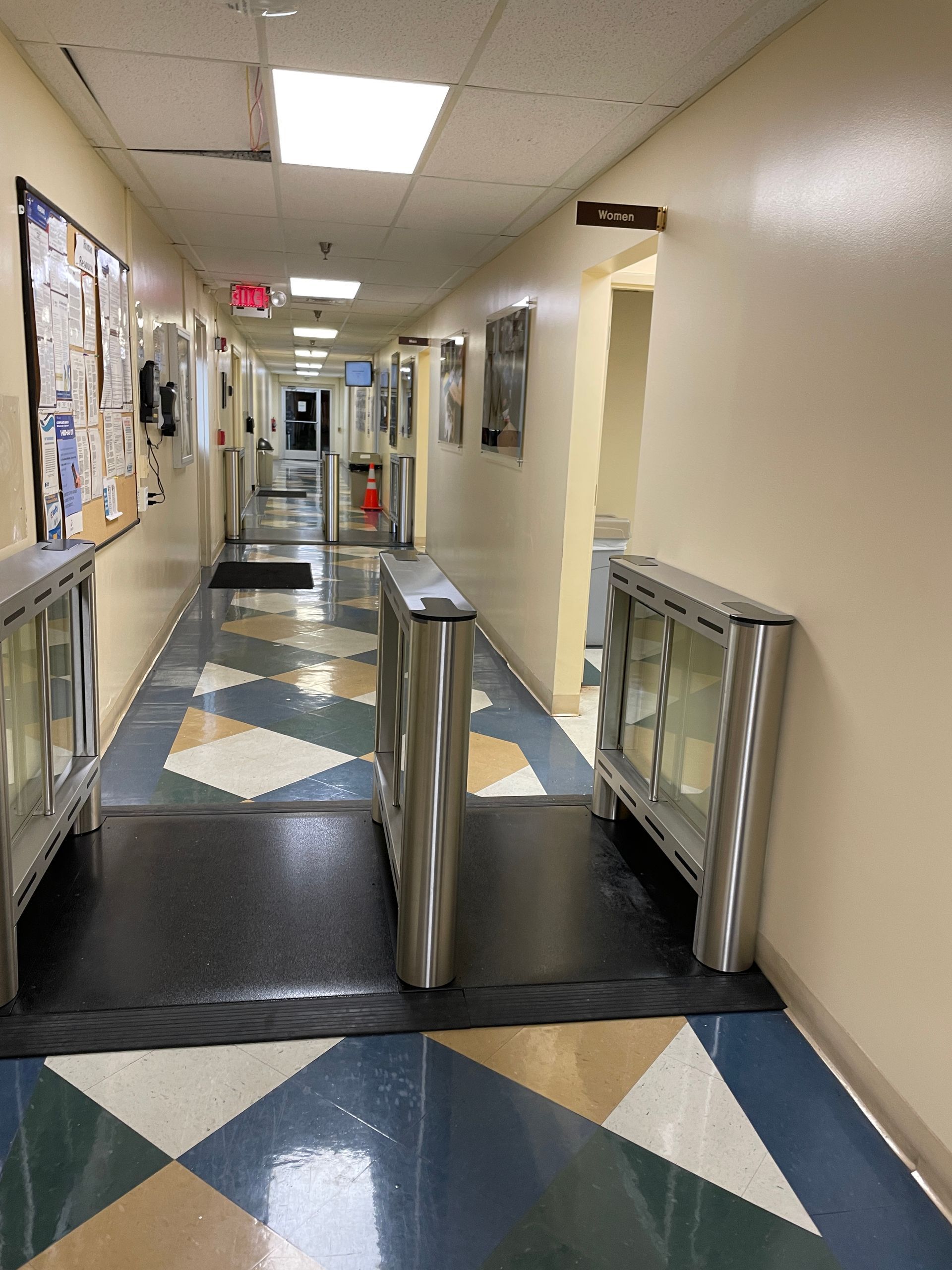Hallway with security turnstiles, patterned flooring, and bulletin board on the left.