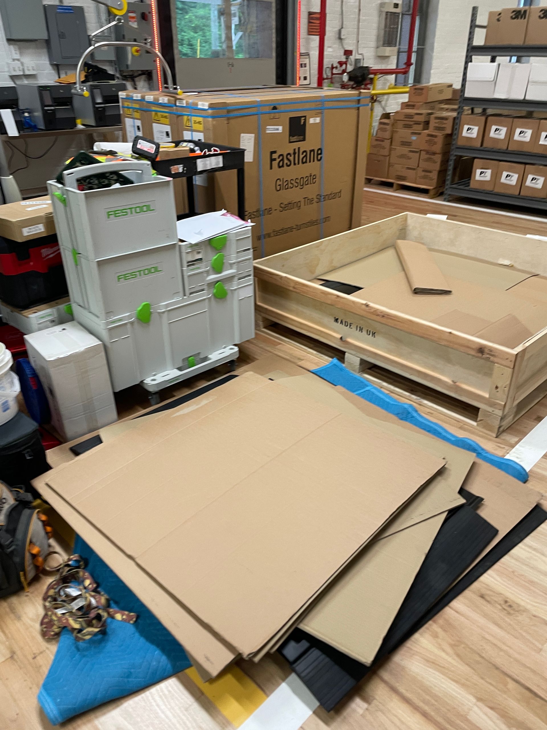 A cluttered warehouse scene with cardboard sheets, wooden crate, plastic bin, and boxed item.
