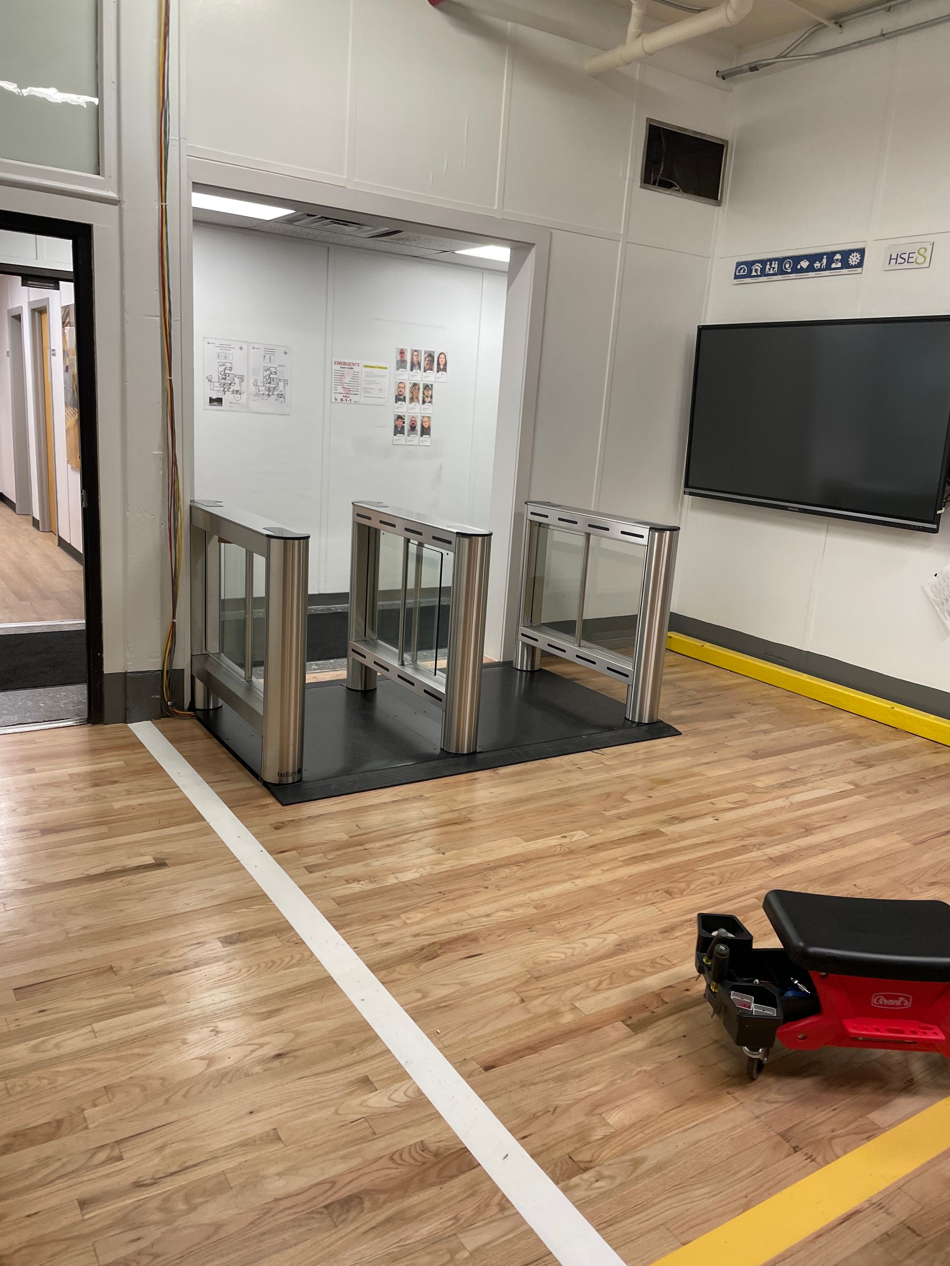 Gym entrance with turnstiles. Interior with wood floor, white lines, a screen, and an open doorway.