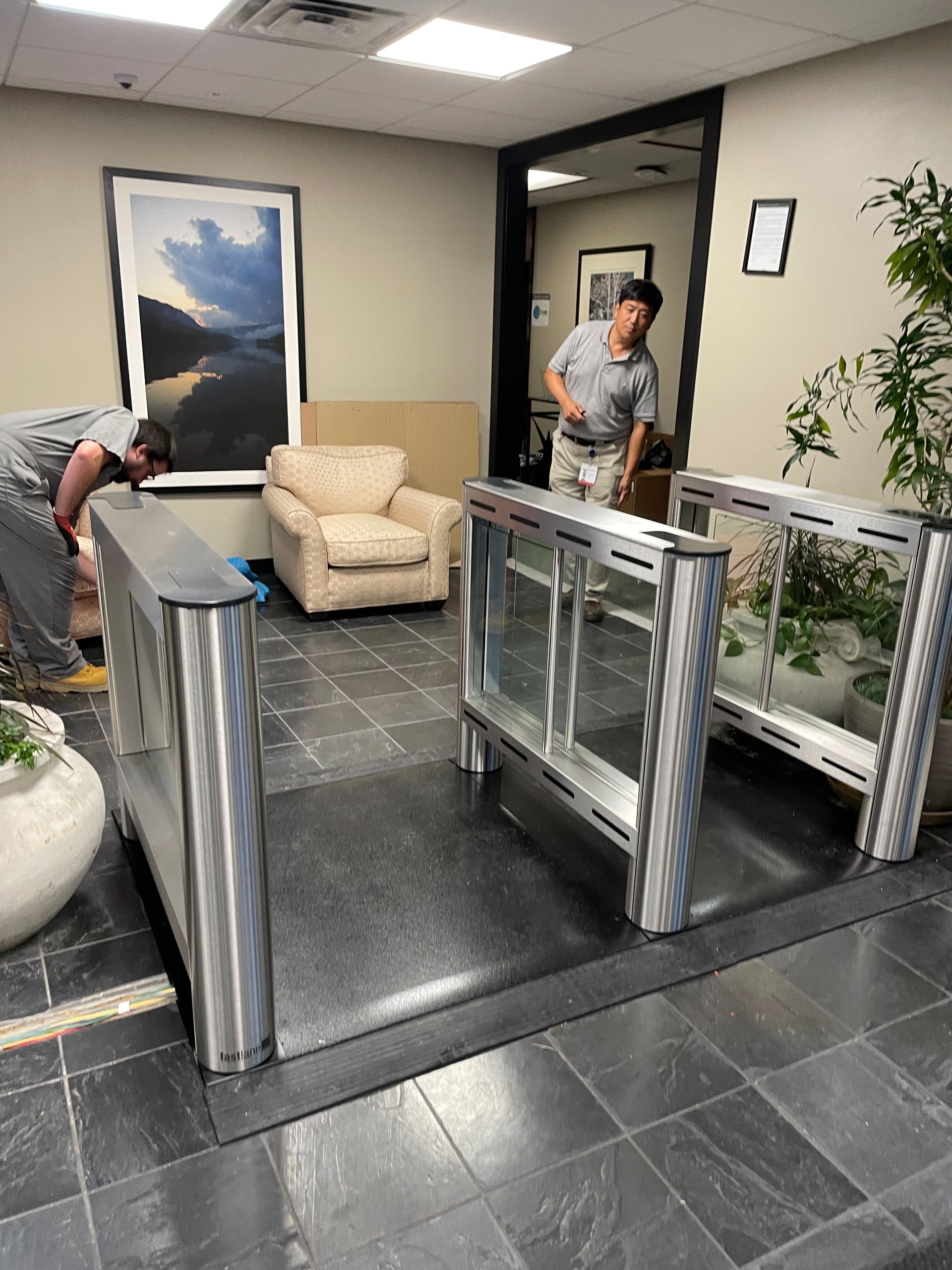 Two people installing security turnstiles in an office lobby with a painting, armchair, and plants.