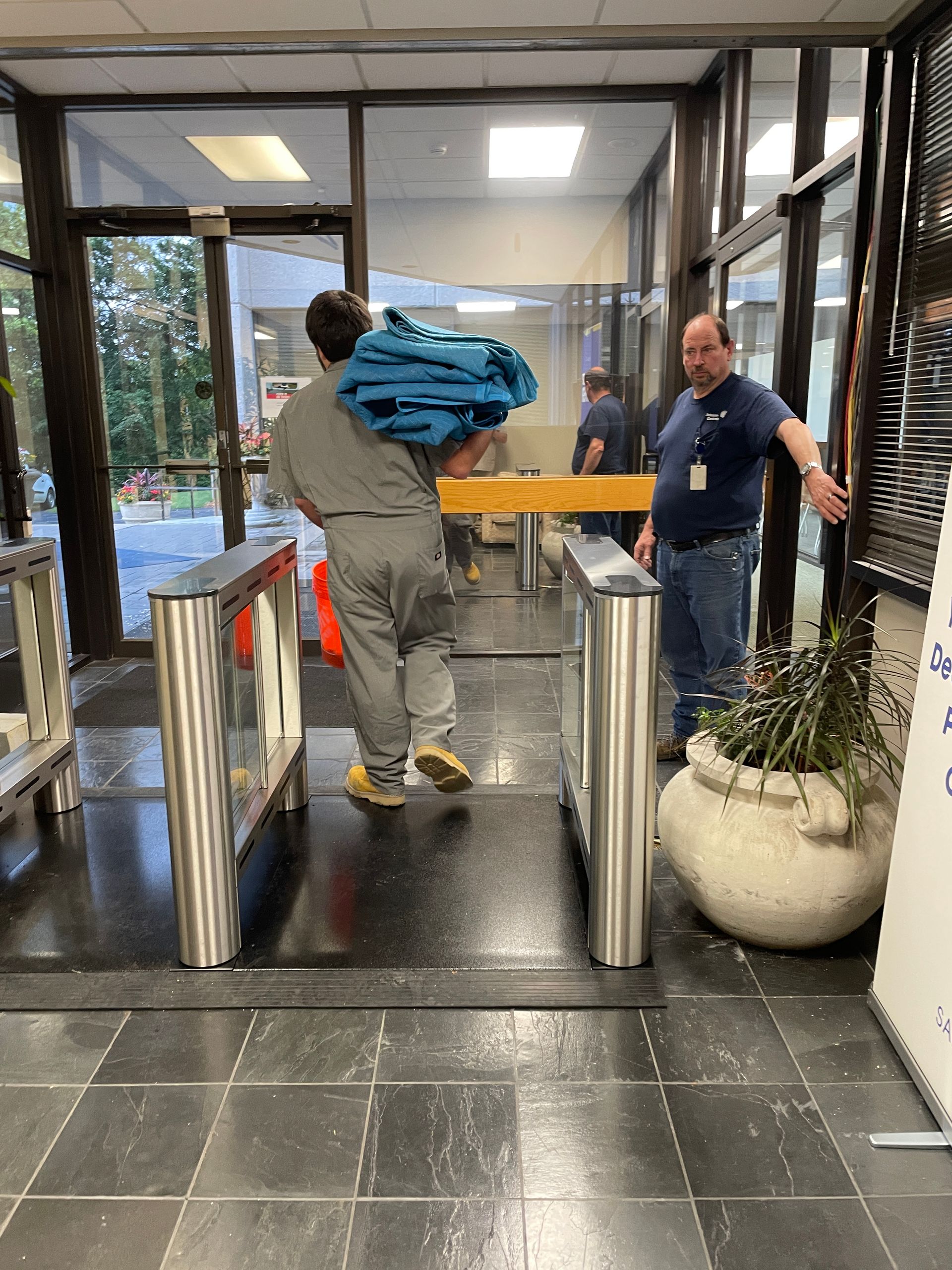 A person carrying blue items walks through security turnstiles as another person points toward the door.