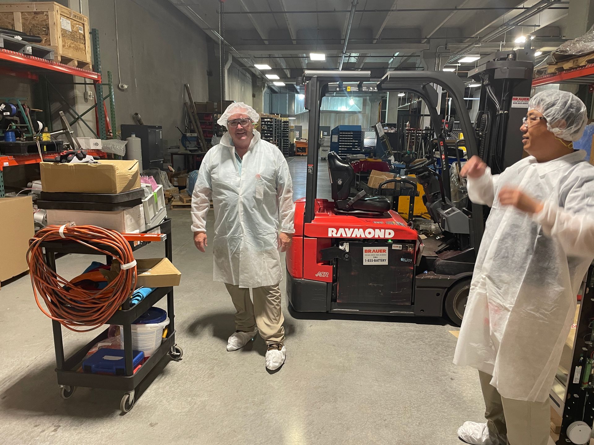 Two people in protective clothing stand in a warehouse with a forklift and equipment.