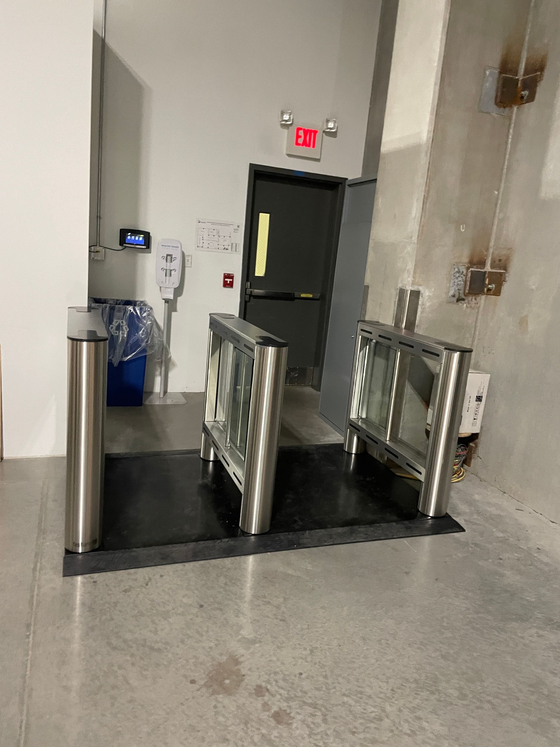 Turnstile entrance with door, exit sign, hand sanitizer, and trash can.